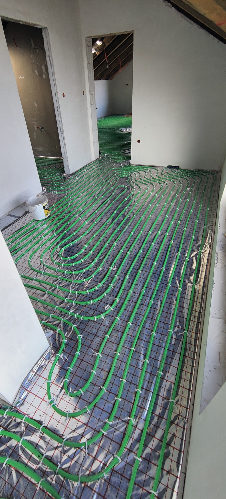 Montaż ogrzewania podłogowego z zielonych rur na folii refleksyjnej w surowym wnętrzu z otwartymi drzwiami do kolejnego pomieszczenia.