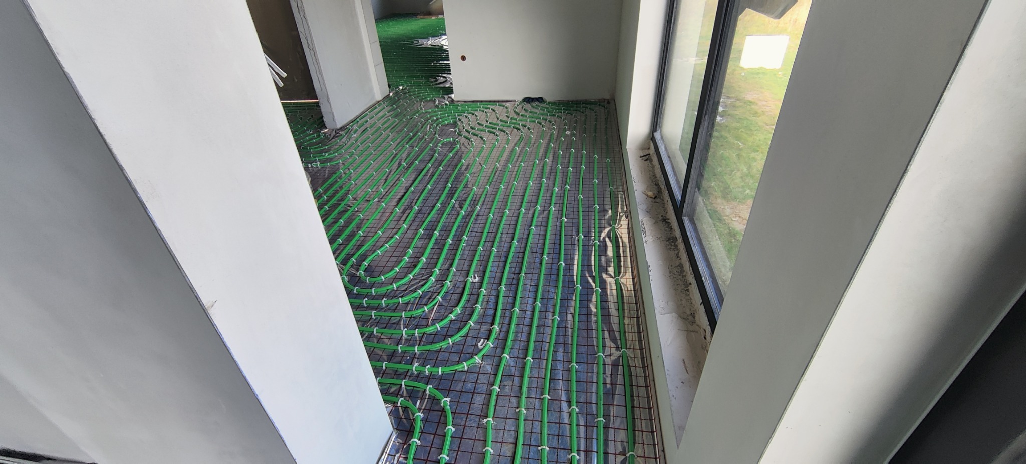 Zielone rurki ogrzewania podłogowego zamocowane do siatki na podłożu w trakcie montażu, widok z góry, surowe wnętrze w trakcie wykańczania.