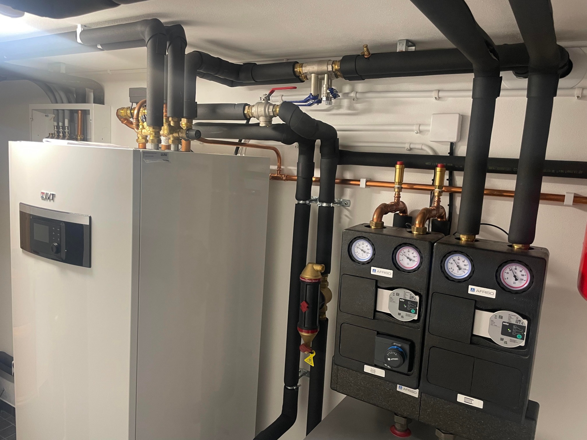 Nowoczesny, biały piec grzewczy z czarnymi, izolowanymi rurami, manometrami i zaworami, zainstalowany w schludnym pomieszczeniu technicznym z białymi ścianami.