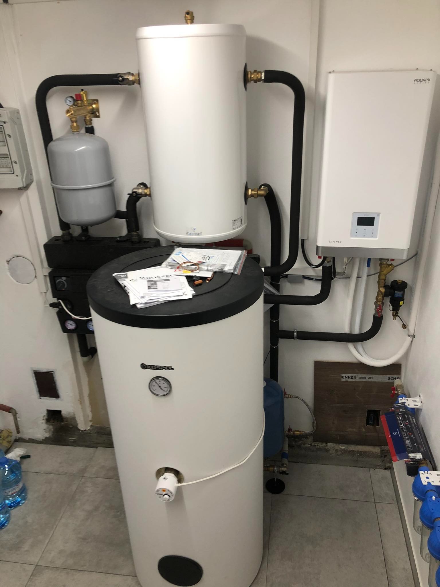 Nowoczesna kotłownia z białym zasobnikiem ciepłej wody, piecem gazowym Rotenso i czarnymi rurami instalacji grzewczej, widoczne dokumenty Kospel na zasobniku.