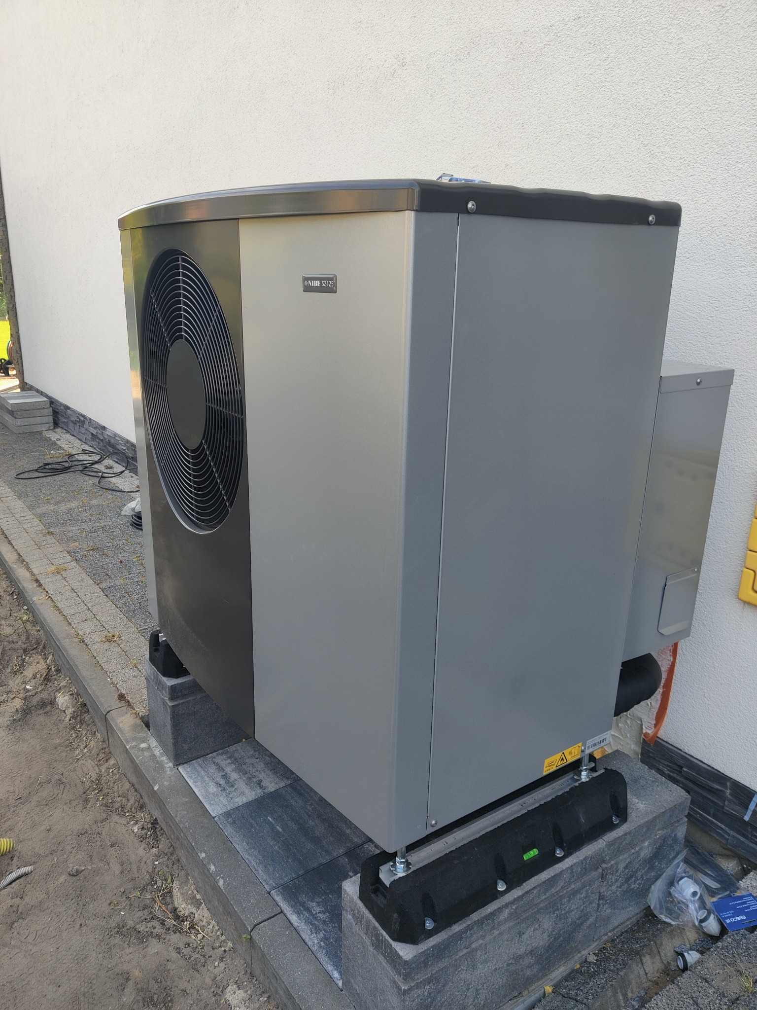 Szara pompa ciepła NIBE S2125 zainstalowana na betonowych bloczkach przy białej ścianie budynku, widoczne elementy instalacji i kostka brukowa.