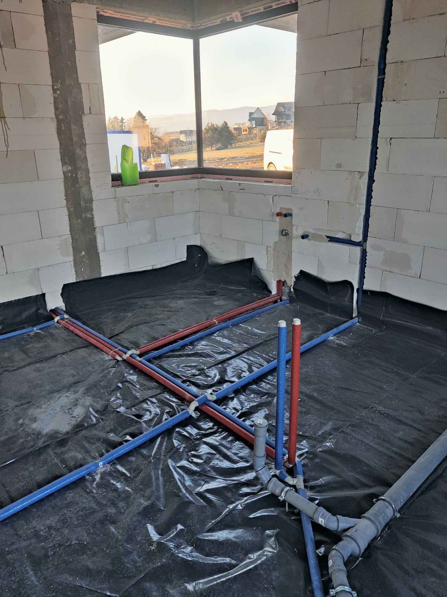 Wnętrze w trakcie prac hydraulicznych: widoczne rury grzewcze i wodne w kolorze czerwonym i niebieskim, rozprowadzone na czarnej folii izolacyjnej, ściany z białego betonu komórkowego, okno...