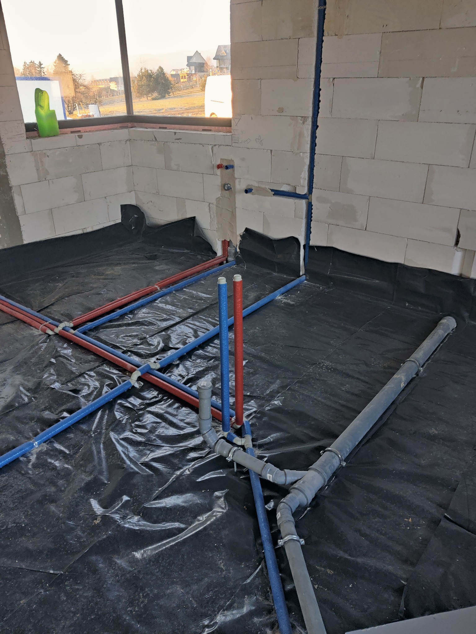 Wnętrze w trakcie budowy z widoczną instalacją wodną i kanalizacyjną: czerwone i niebieskie rury PEX krzyżują się na czarnej folii, pionowa niebieska rura przy ścianie z betonu komórkowego, szara...