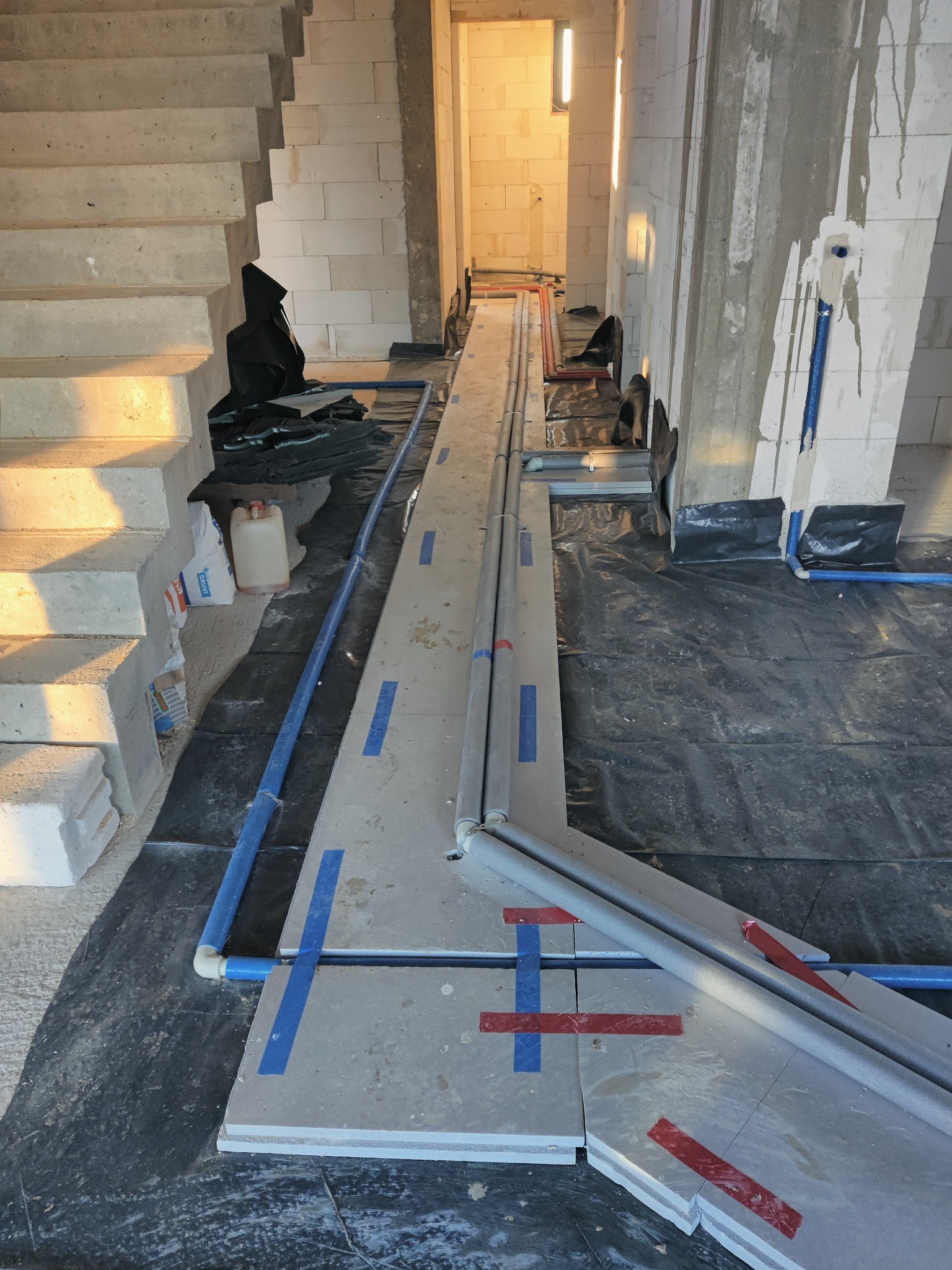 Instalacja ogrzewania podłogowego w trakcie montażu: widoczne rury grzewcze ułożone na izolacji, zabezpieczone taśmą, na tle surowego wnętrza w trakcie budowy ze schodami i ścianami z betonu...