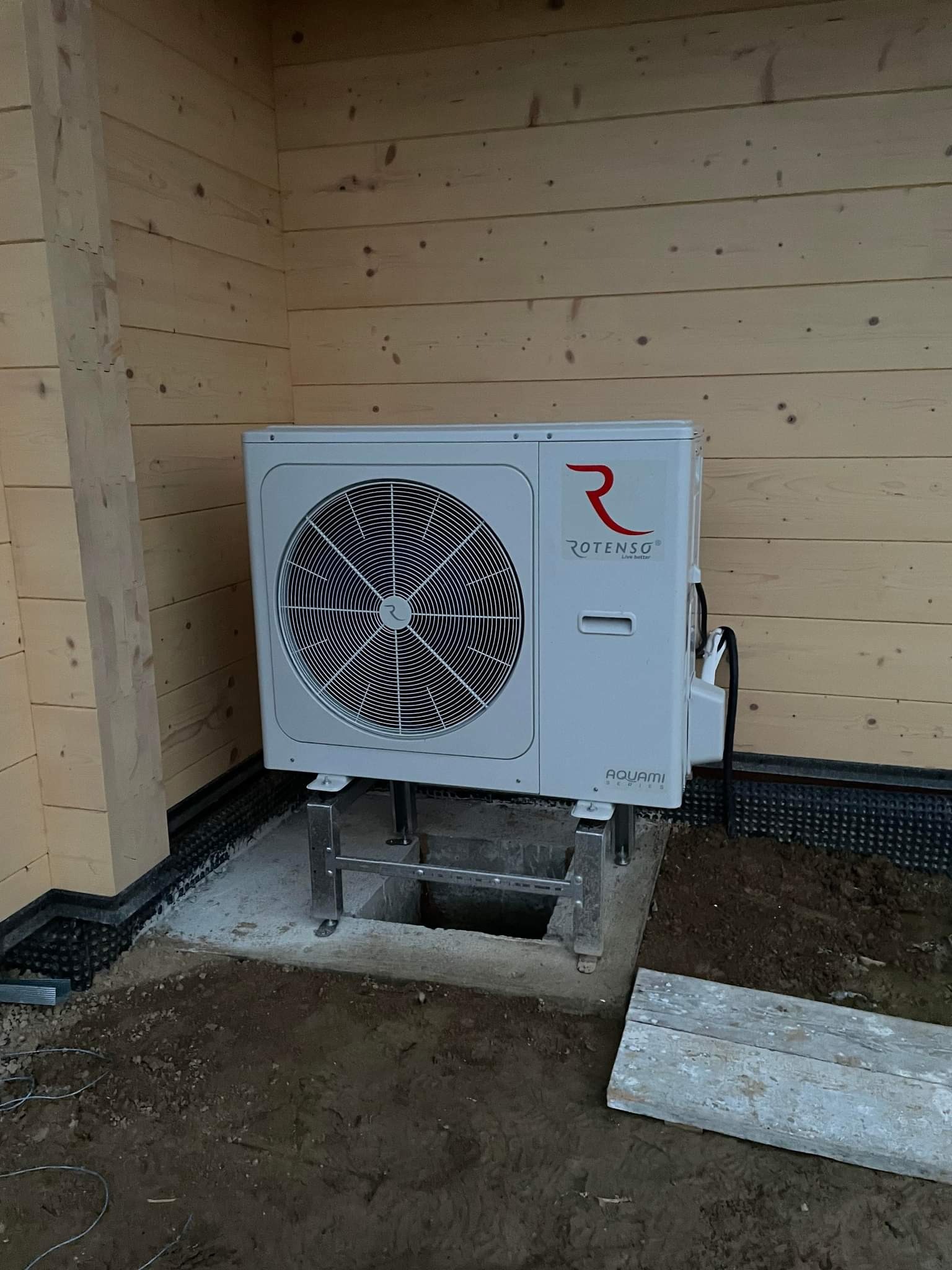 Zewnętrzna jednostka pompy ciepła Rotenso Aquami, zamontowana na metalowym stelażu przy drewnianej elewacji budynku, widoczny otwór w fundamencie.