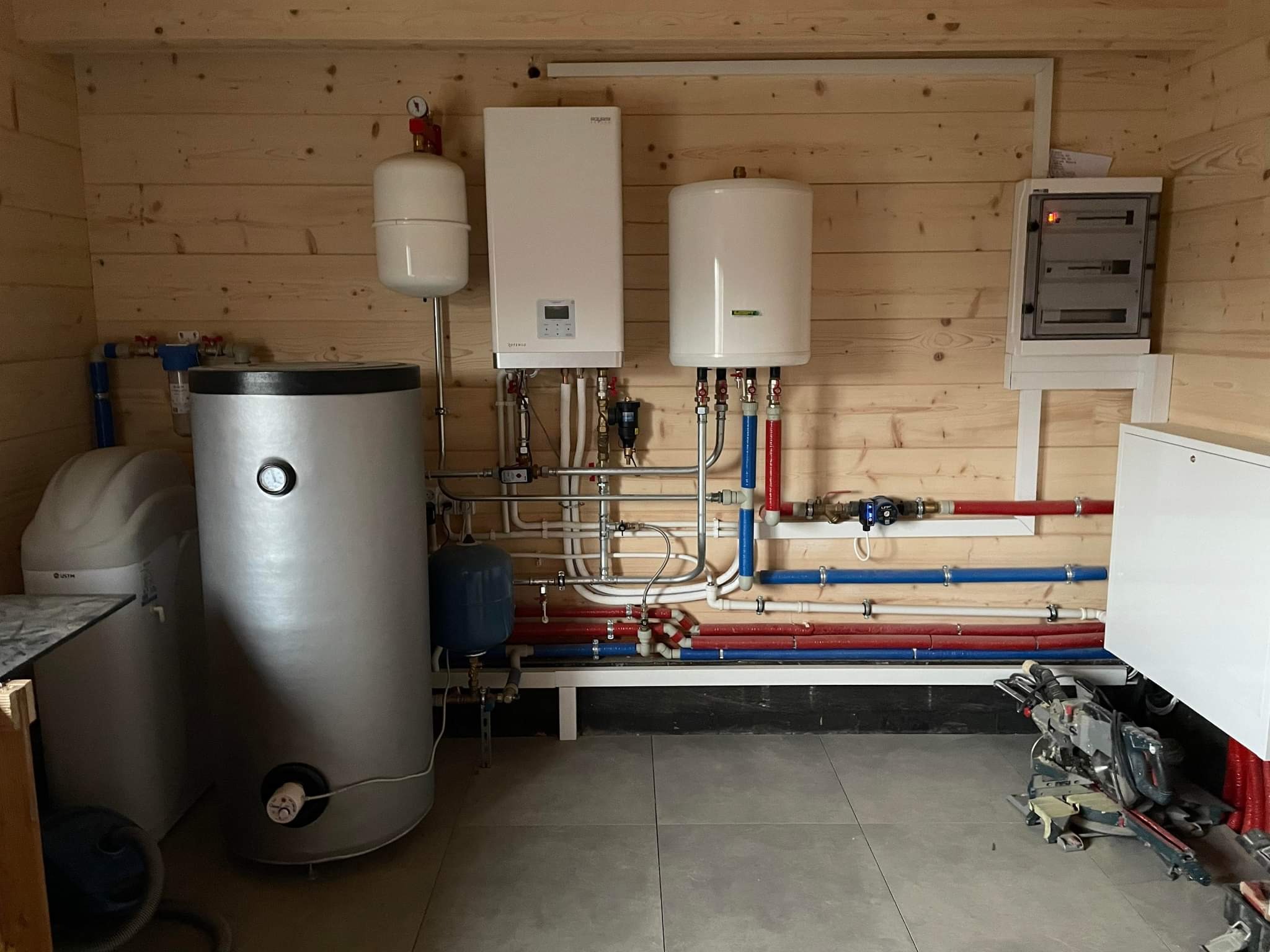 Wnętrze kotłowni wykończonej boazerią, z zainstalowanym kotłem gazowym, zasobnikiem ciepłej wody, naczyniami przeponowymi, zmiękczaczem wody i rozdzielnicą elektryczną, widoczne rury instalacji...