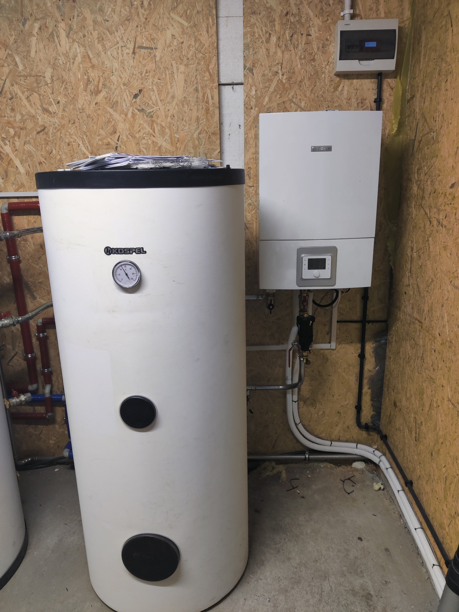 Biały zasobnik ciepłej wody użytkowej marki Kospel z analogowym termometrem, obok wiszący piec gazowy Bosch z cyfrowym wyświetlaczem, zamontowane na ścianie z płyty OSB, z widoczną instalacją...