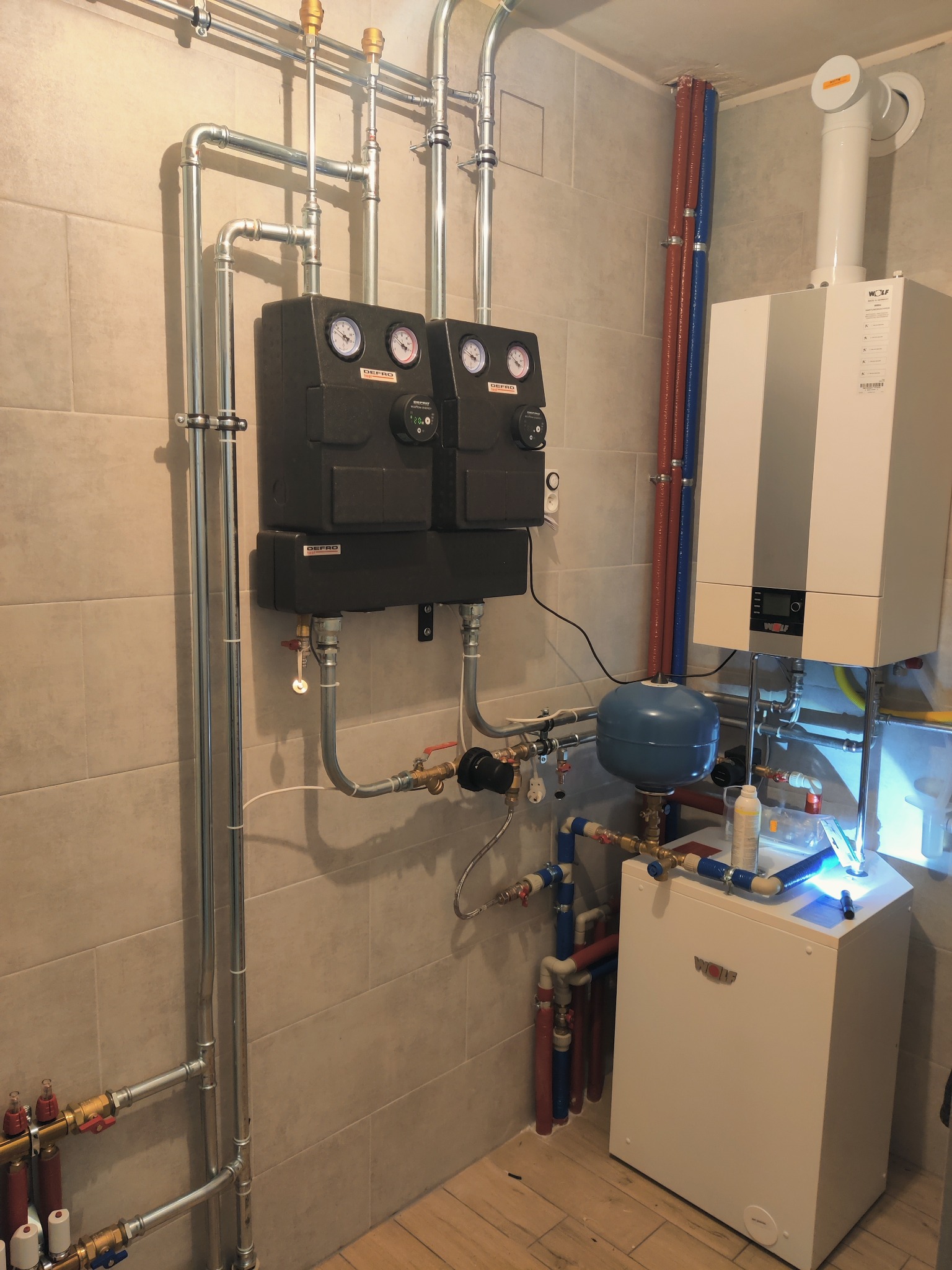 Złożona instalacja grzewcza w kotłowni z piecem gazowym Wolf, rozdzielaczami Defro z manometrami i licznikiem ciepła, oraz rurami w kolorach niebieskim i czerwonym, widoczne na tle płytek...