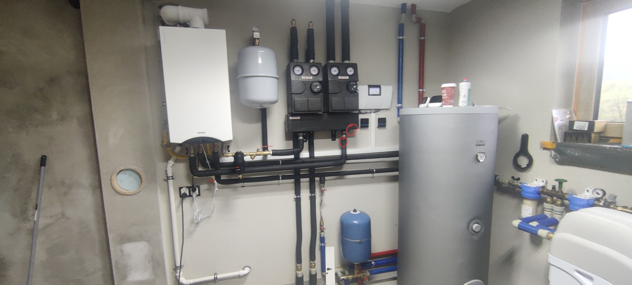 Złożona instalacja grzewcza z piecem gazowym, zbiornikiem i rozdzielnicą, rury w izolacji, manometry, filtry, widoczne elementy sterowania. Szara ściana w tle.