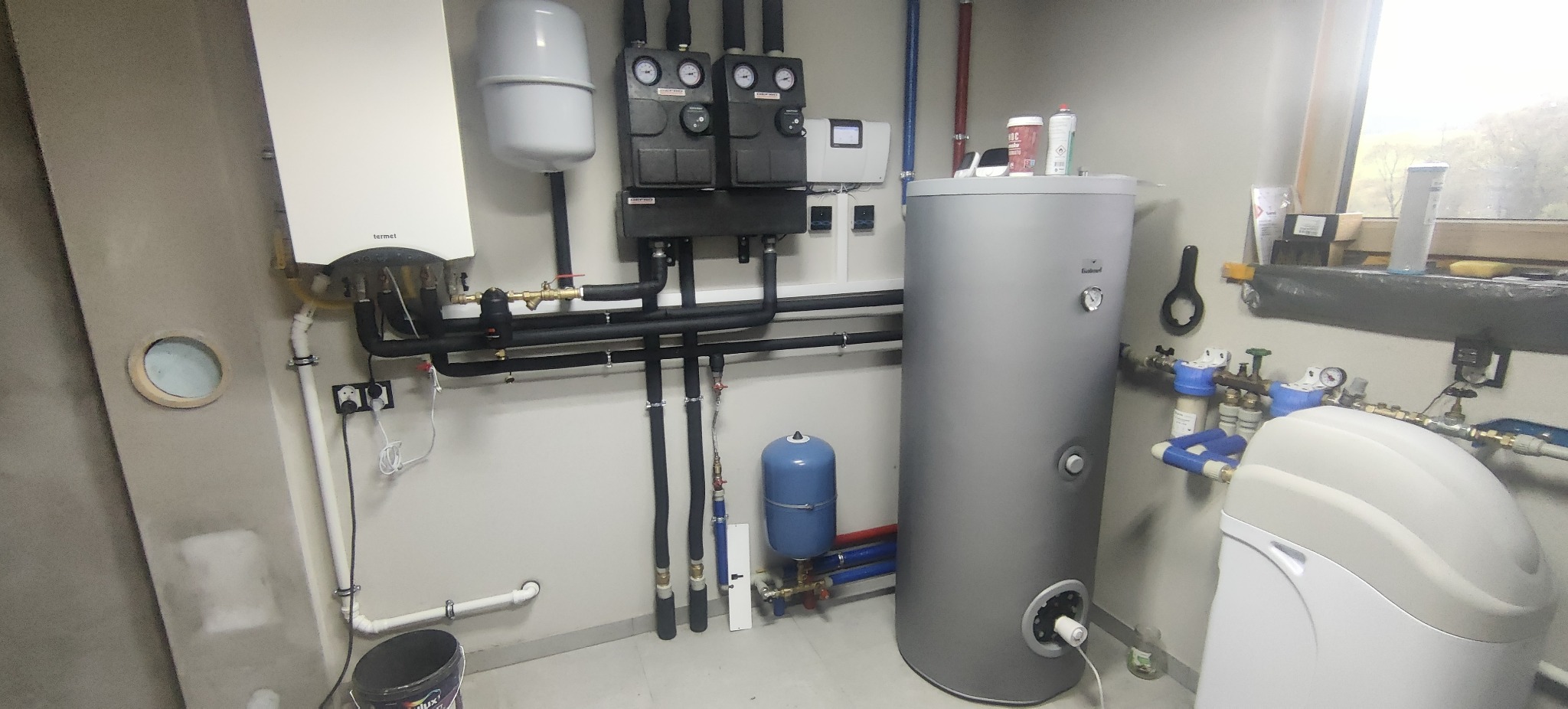 Zestaw urządzeń grzewczych w kotłowni: piec gazowy Termet, zbiornik Galmet, naczynia przeponowe, filtry i armatura. Widoczne rury w izolacji i okno w tle.