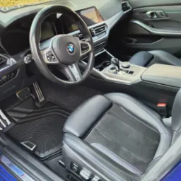 Wnętrze samochodu BMW serii 3 z czarną tapicerką z alcantary i niebieskimi przeszyciami, widoczne dywaniki gumowe z logo BMW M Performance oraz aluminiowe pedały.
