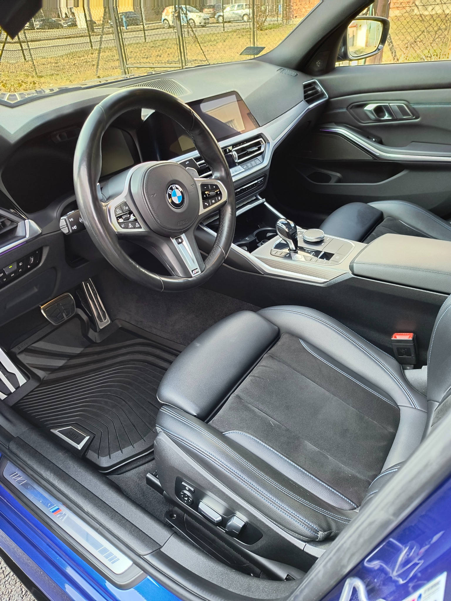 Wnętrze samochodu BMW serii 3 z czarną tapicerką z alcantary i niebieskimi przeszyciami, widoczne dywaniki gumowe z logo BMW M Performance oraz aluminiowe pedały.
