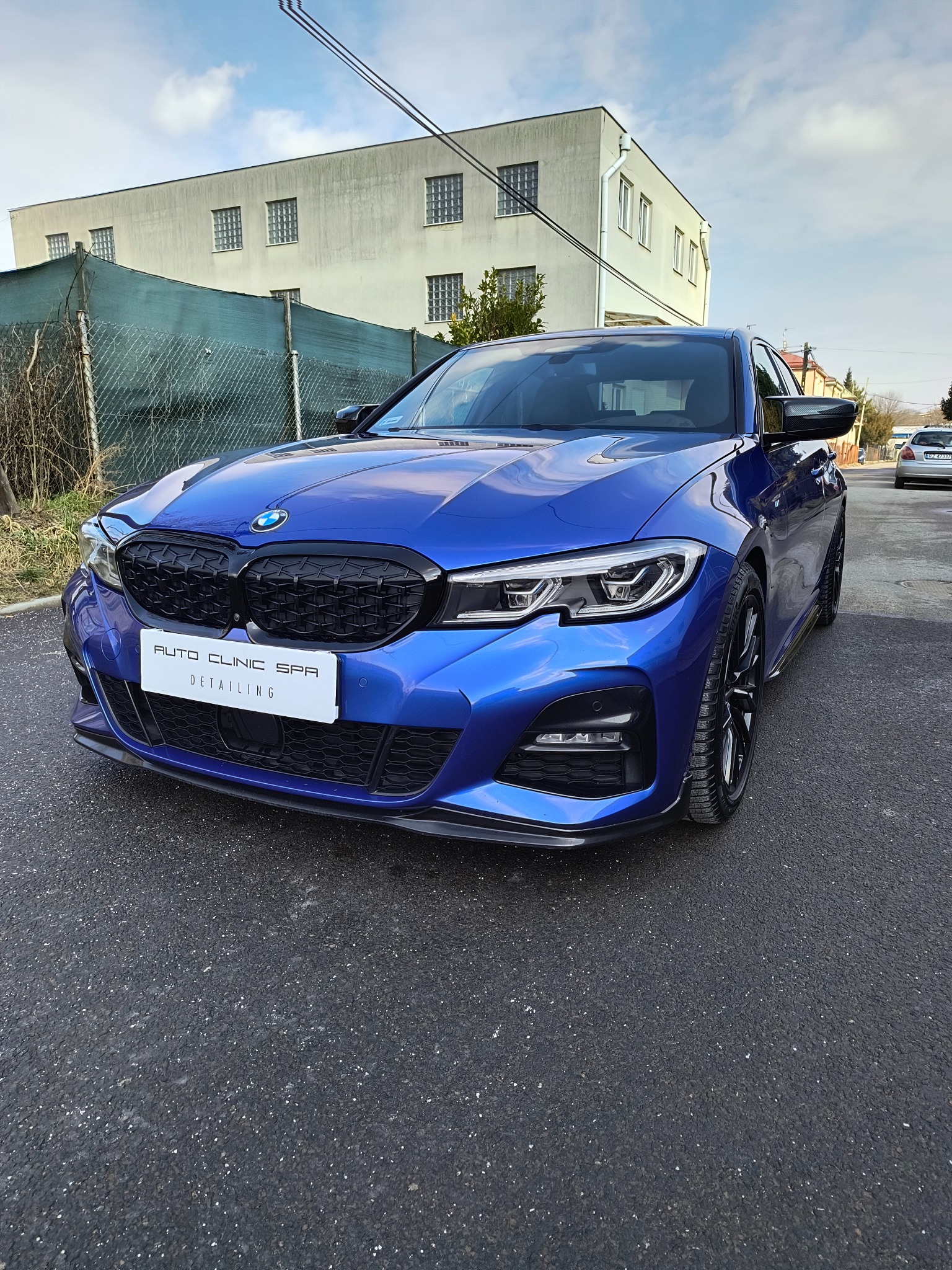 Błękitny samochód BMW z czarnym grillem i tablicą 'Auto Clinic Spa Detailing' na tle budynku i pochmurnego nieba.