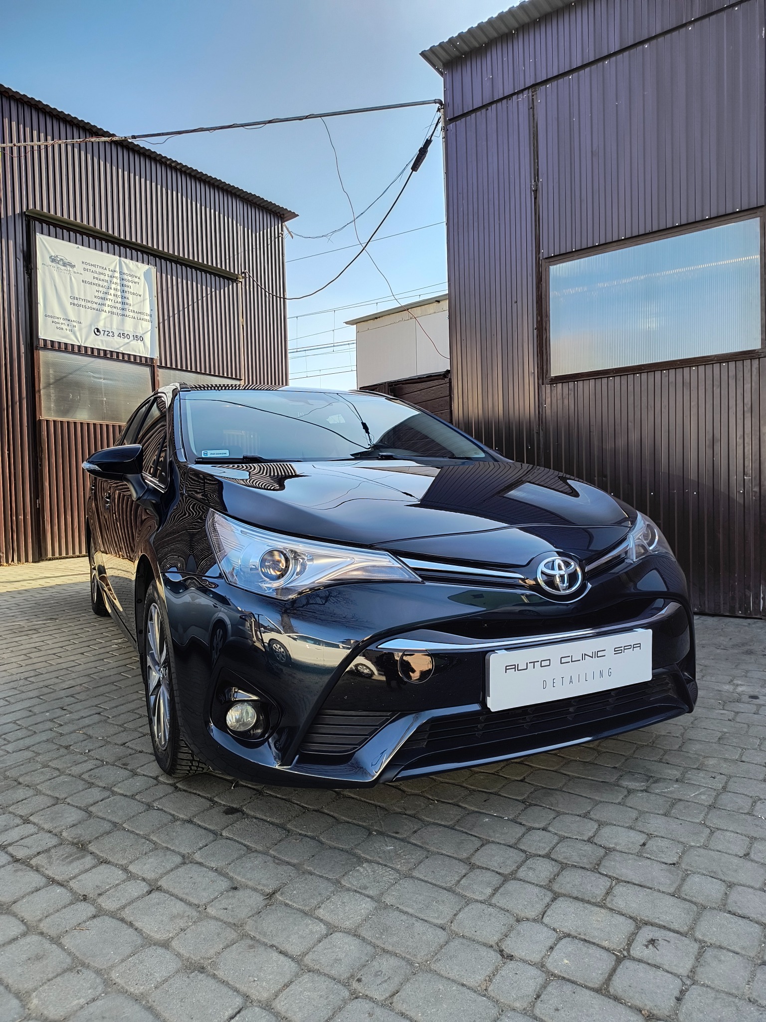 Błyszczący, czarny samochód Toyota Avensis zaparkowany na kostce brukowej przed budynkiem myjni samochodowej Auto Clinic Spa Detailing. Widoczna tabliczka z ofertą usług i numerem telefonu.