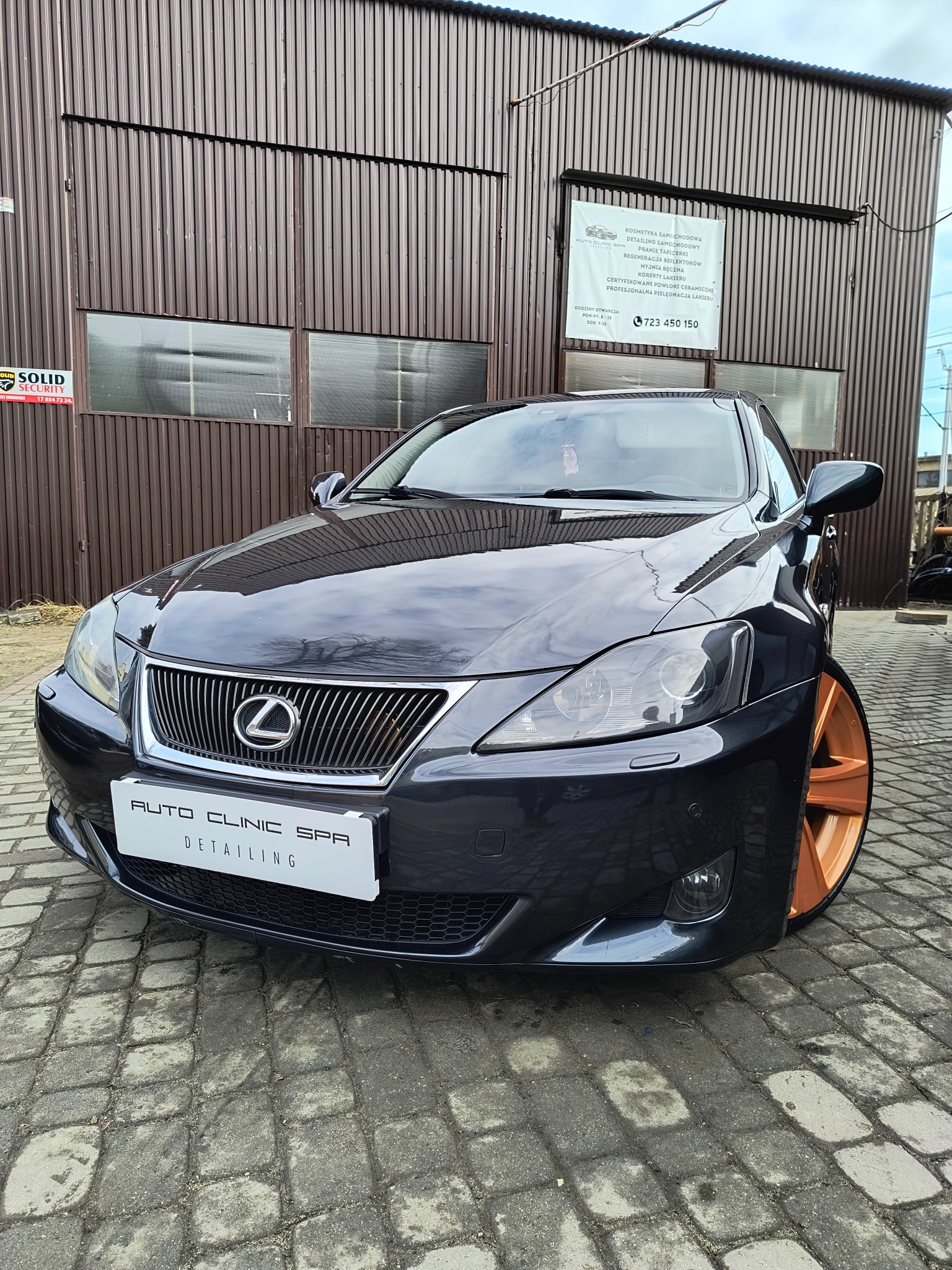Czarny Lexus IS z pomarańczowymi felgami zaparkowany przed budynkiem z szyldem 'Auto Clinic Spa Detailing' oferującym kosmetykę samochodową, pranie tapicerki i myjnię ręczną.