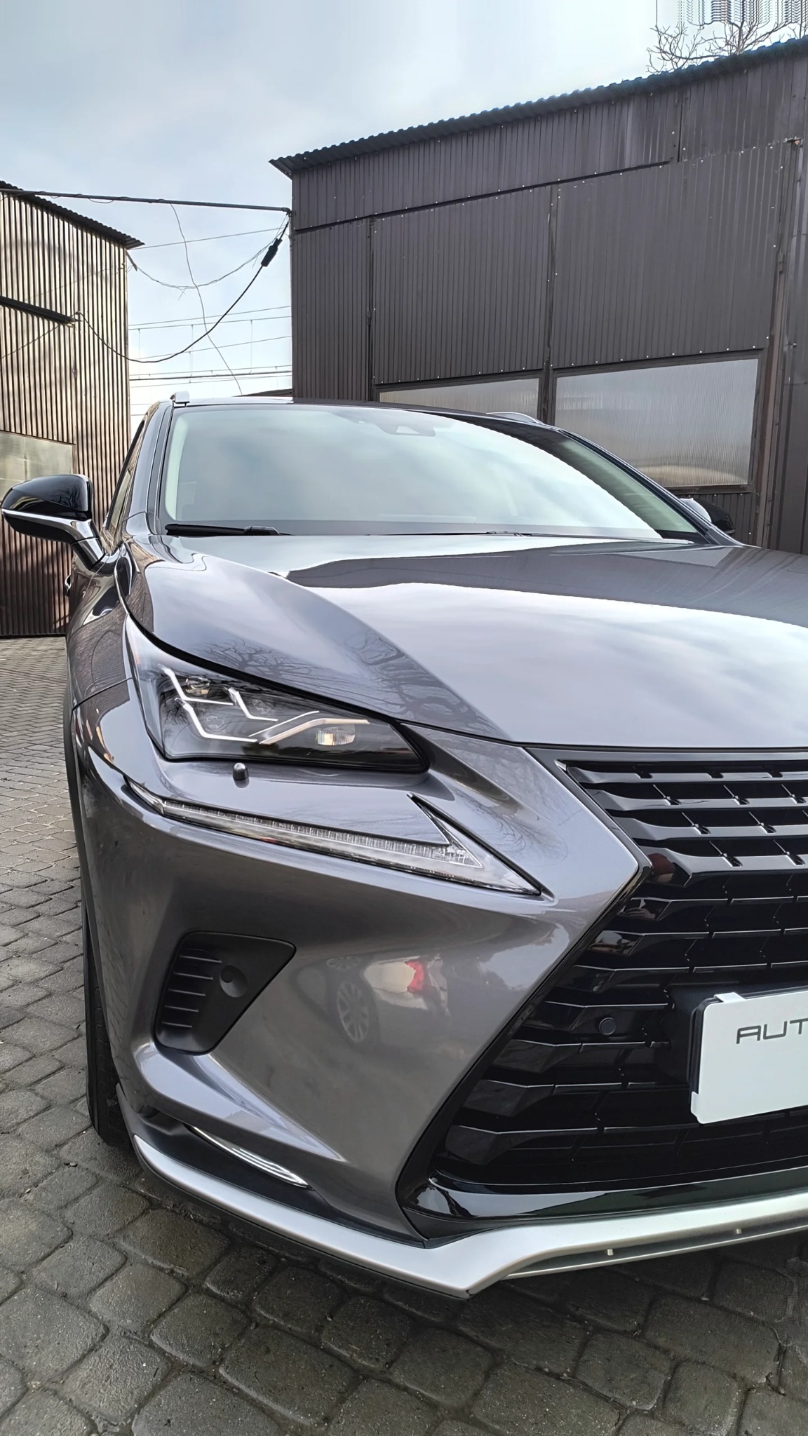 Szary samochód Lexus na tle blaszanych garaży, widok z przodu na reflektor i grill.