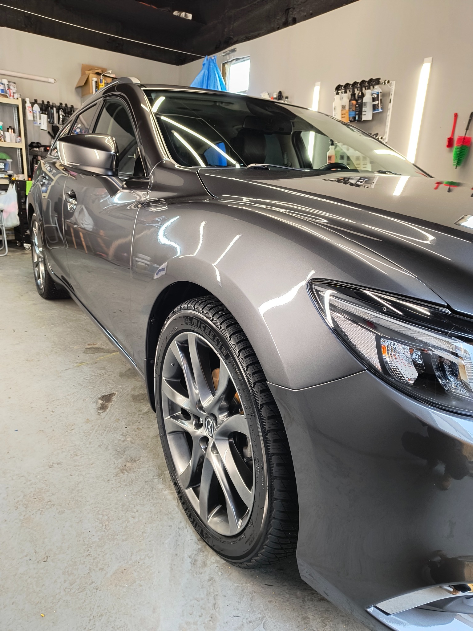 Błyszczący, szary samochód po umyciu, widoczne krople wody na karoserii, opona z wyraźnym bieżnikiem i felga z logo producenta, w tle półki z preparatami do pielęgnacji aut.