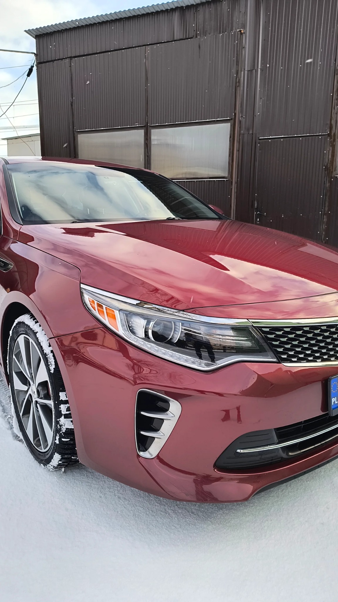 Bordowy samochód marki Kia Optima, częściowo pokryty śniegiem, zaparkowany przed ciemnobrązowym budynkiem z metalowymi panelami.