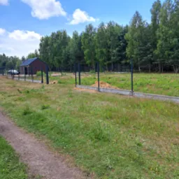 Fundament pod ogrodzenie z zamontowanymi słupkami na zalesionej działce, widoczny pryzmat piasku i budynek w tle.