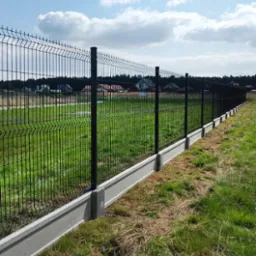 Długi, czarny płot z siatki na betonowej podmur&oacute;wce, chroniący posesję z zielonym trawnikiem i domami w tle, widok z boku.