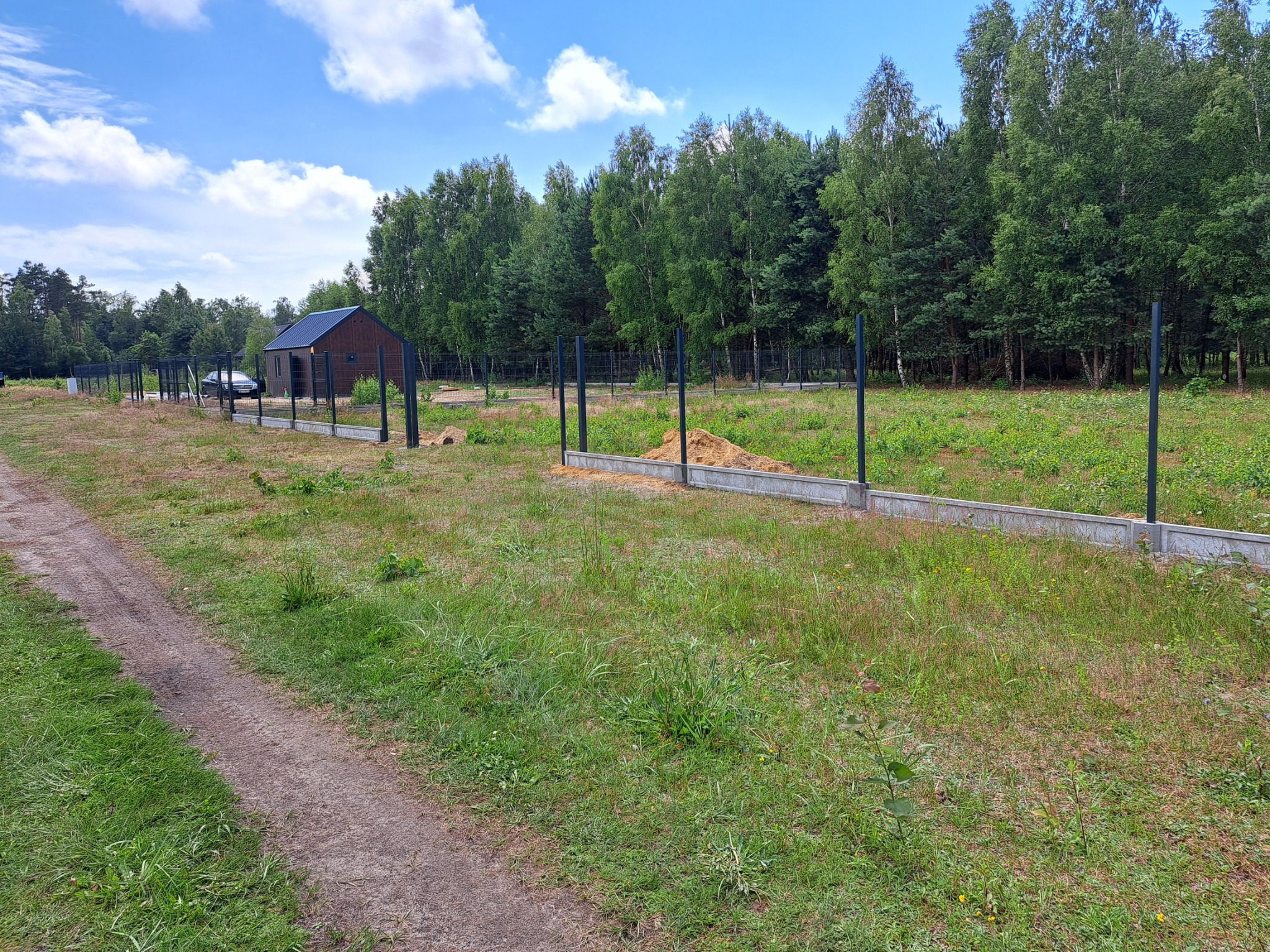 Fundament pod ogrodzenie z zamontowanymi słupkami na zalesionej działce, widoczny pryzmat piasku i budynek w tle.