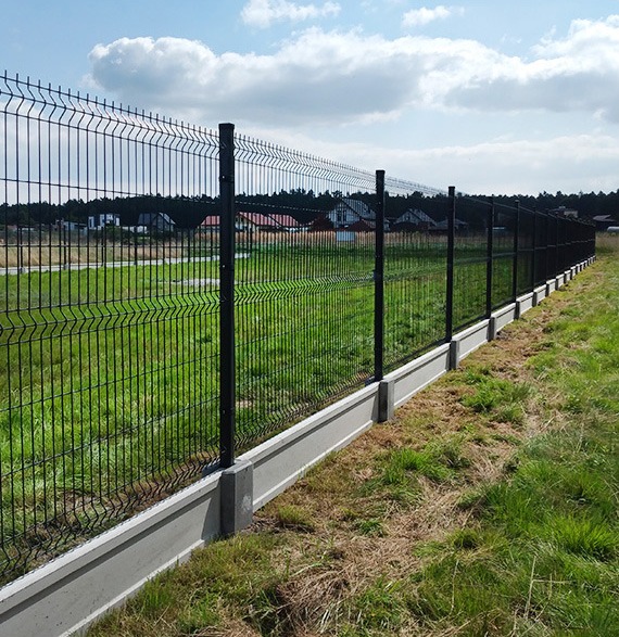 Długi, czarny płot z siatki na betonowej podmurówce, chroniący posesję z zielonym trawnikiem i domami w tle, widok z boku.