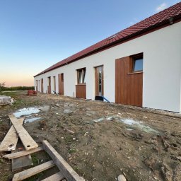 Maksym Kolosovskyi - Długi parterowy budynek z białą elewacją i brązowymi akcentami, w trakcie prac wykończeniowych, widoczne błoto i materiały budowlane na pierwszym planie.