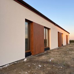 Maksym Kolosovskyi - Nowoczesny dom jednorodzinny z jasną elewacją i drewnianymi okładzinami okiennymi, widoczne prace wykończeniowe wokół budynku, ziemia w stanie surowym, ciepłe światło zachodzącego słońca.