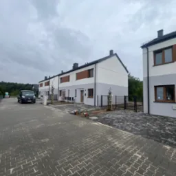 Szeregówka domów jednorodzinnych z elewacją w odcieniach bieli i szarości, z brązowymi akcentami, widok z ulicy wyłożonej kostką, pochmurne niebo.