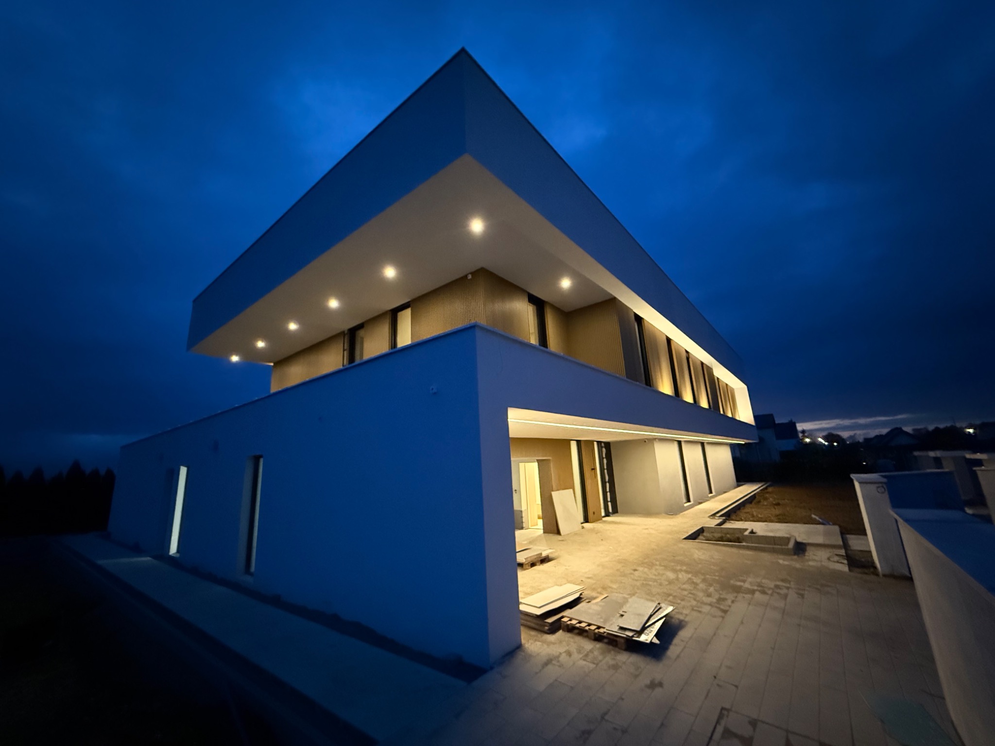 Nowoczesny, dwukondygnacyjny dom z płaskim dachem oświetlony punktowymi lampami w elewacji, sfotografowany o zmierzchu z niskiej perspektywy, ukazujący fragment placu budowy z paletami i płytami.