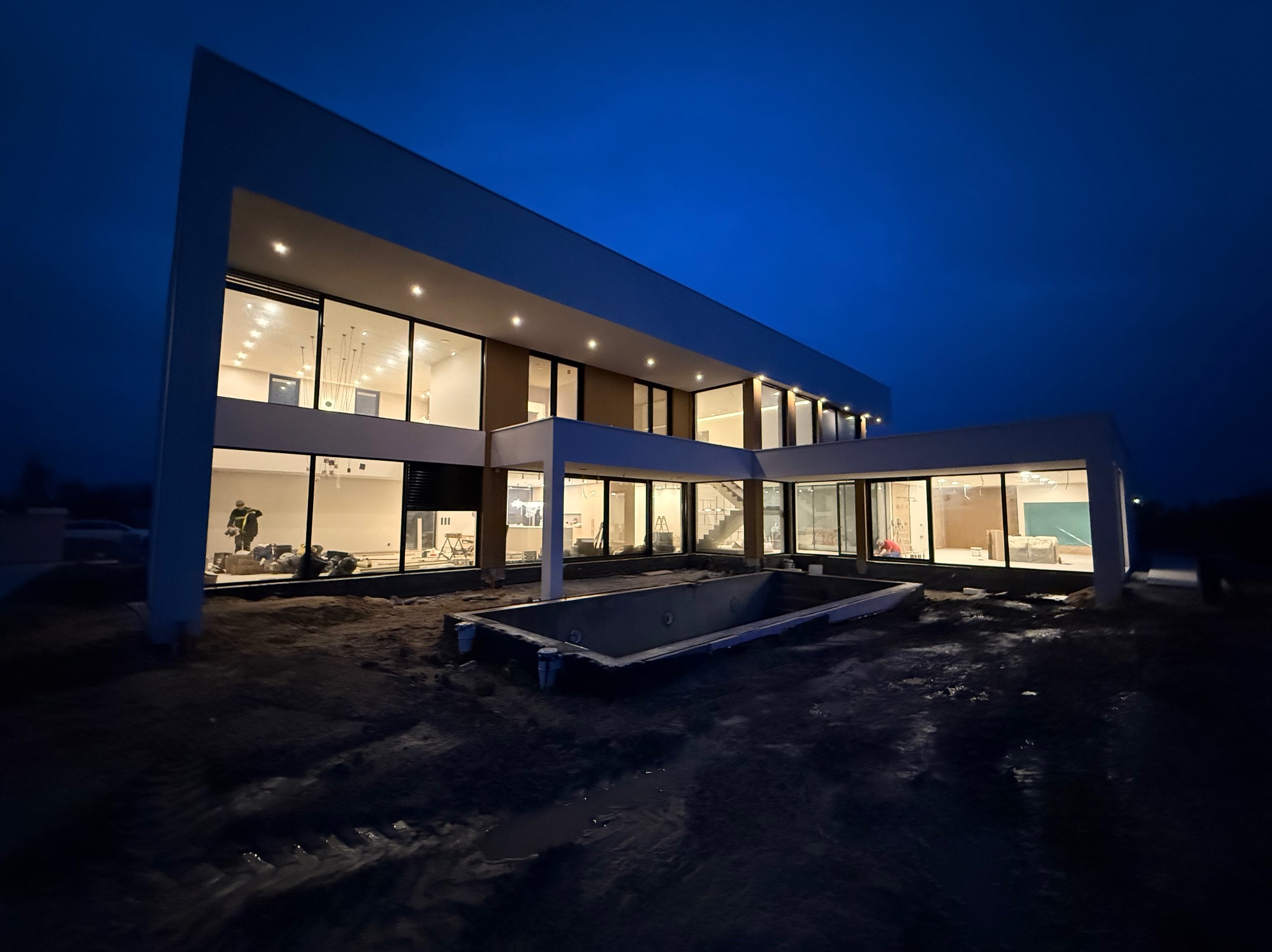 Nowoczesny dom z dużymi oknami i basenem w trakcie budowy o zmierzchu, widoczne prace wykończeniowe wewnątrz.