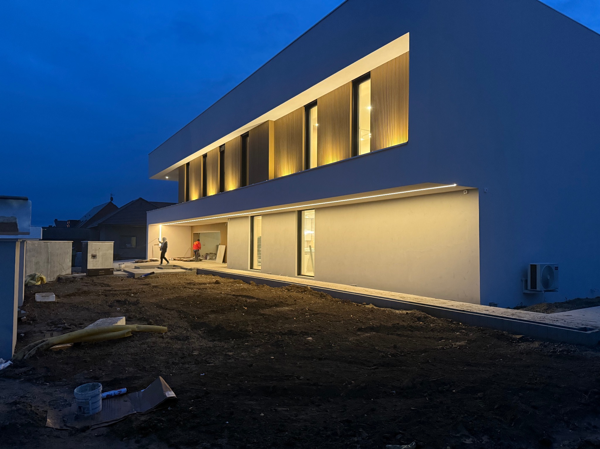 Nowoczesny dom jednorodzinny z oświetloną elewacją w odcieniach bieli i drewna, widoczny podczas zmierzchu, z dwoma pracownikami na placu budowy i niezagospodarowanym terenem wokół.