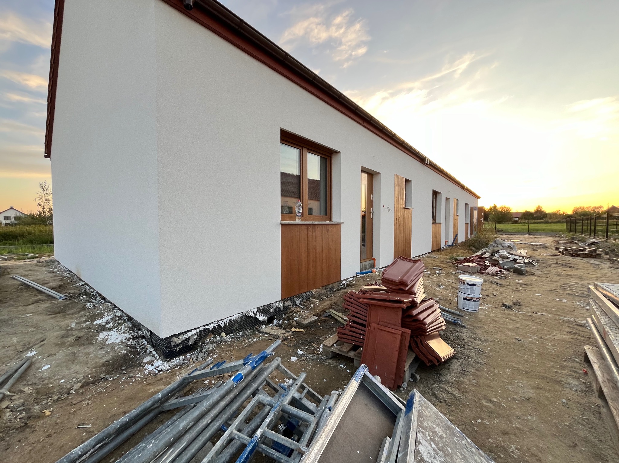 Nowo otynkowany budynek parterowy z brązowymi elementami wykończeniowymi, widok z zewnątrz, w tle zachodzące słońce, na pierwszym planie porozrzucane materiały budowlane i rusztowania.