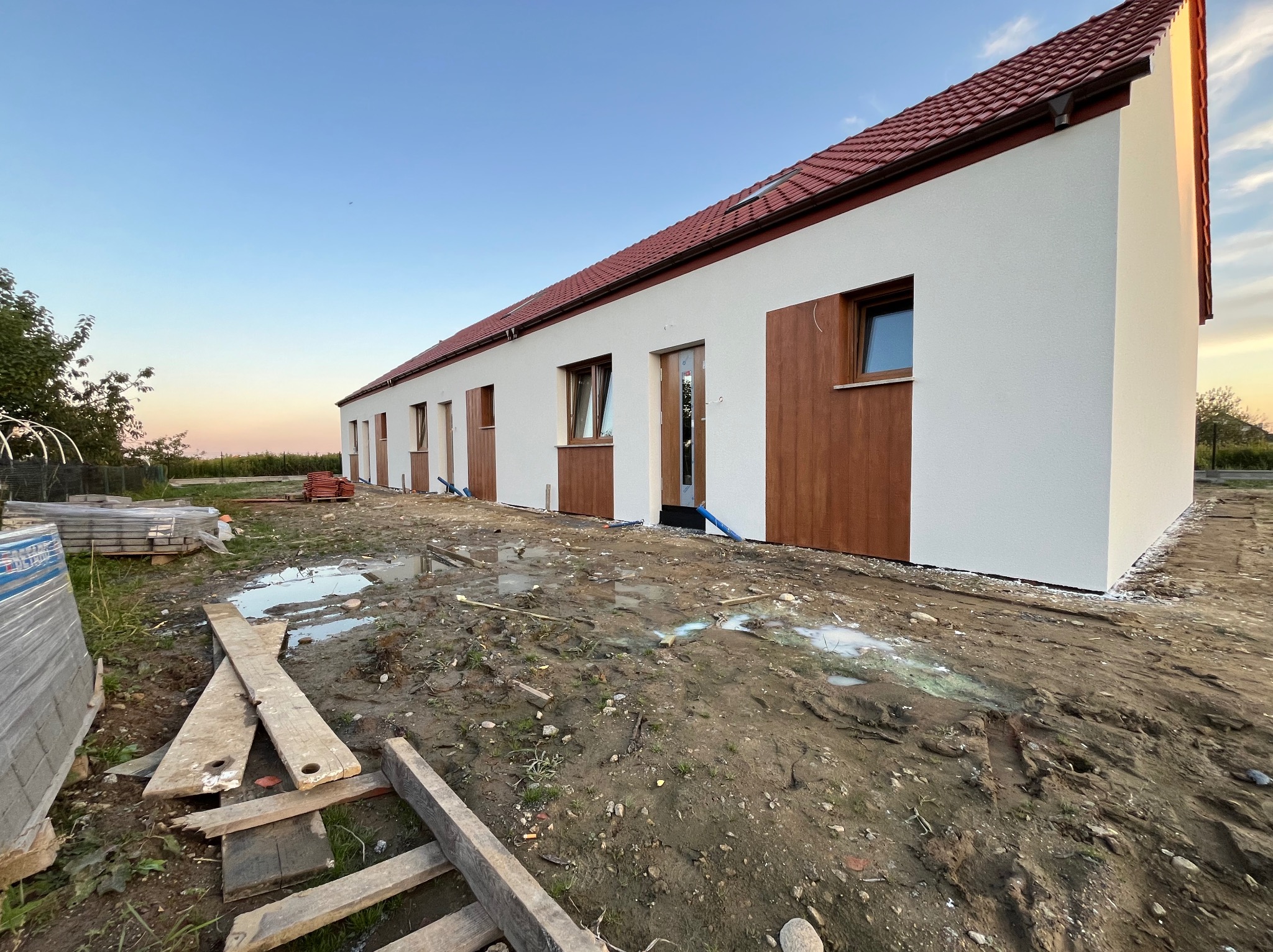 Długi parterowy budynek z białą elewacją i brązowymi akcentami, w trakcie prac wykończeniowych, widoczne błoto i materiały budowlane na pierwszym planie.