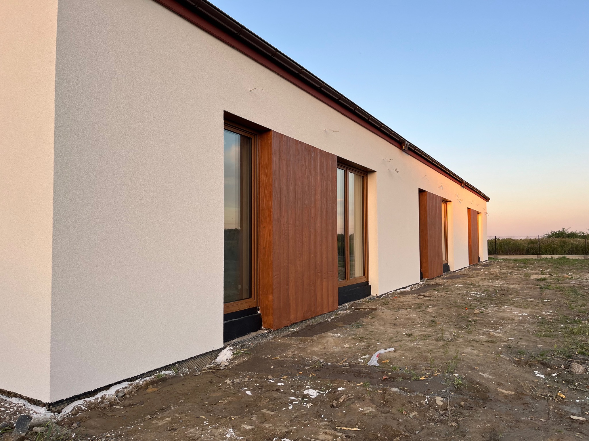 Nowoczesny dom jednorodzinny z jasną elewacją i drewnianymi okładzinami okiennymi, widoczne prace wykończeniowe wokół budynku, ziemia w stanie surowym, ciepłe światło zachodzącego słońca.