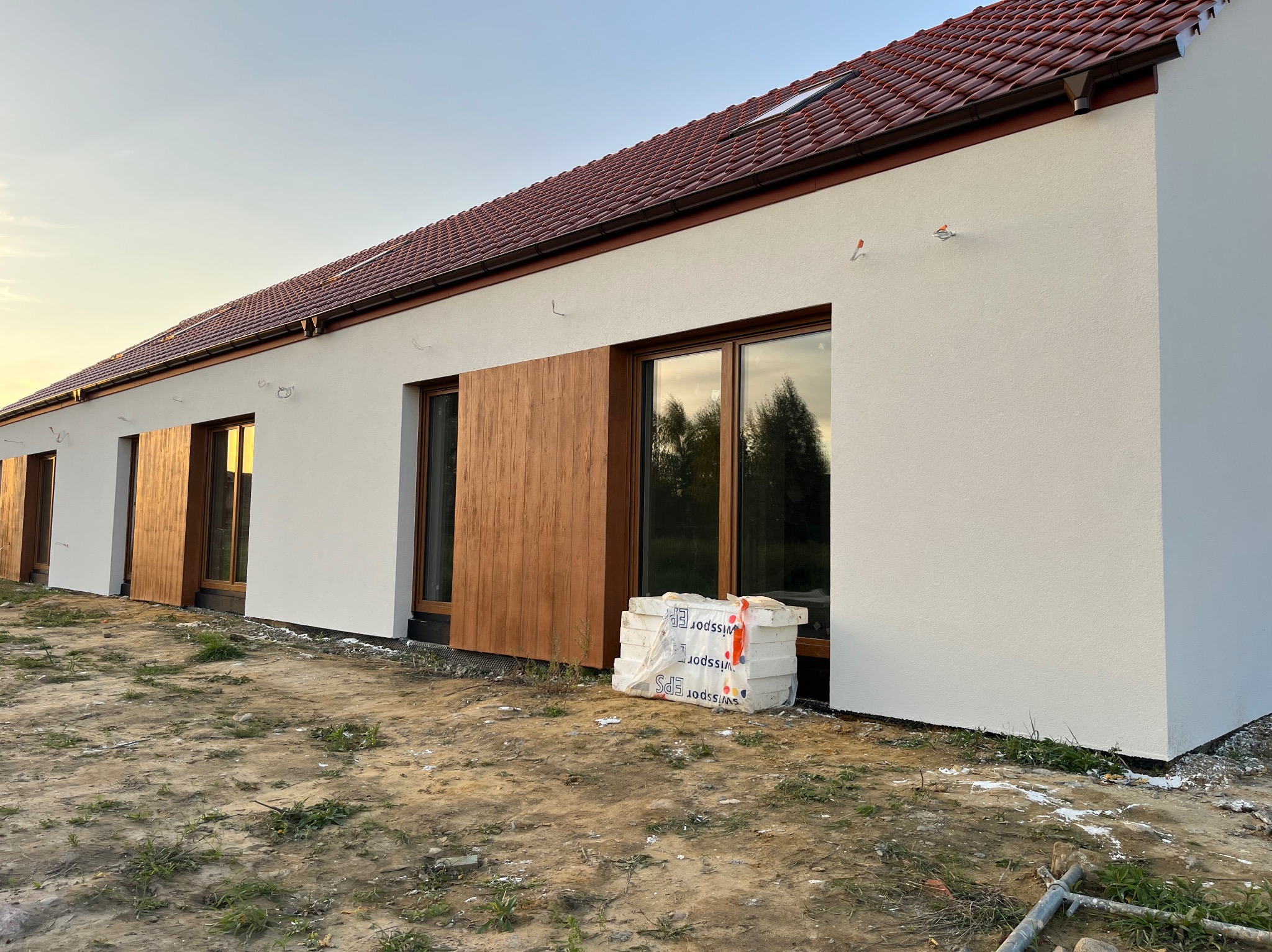 Nowoczesny dom jednorodzinny z jasną elewacją i drewnianymi okiennicami, na podwórku leżą paczki styropianu Swissport EPS.