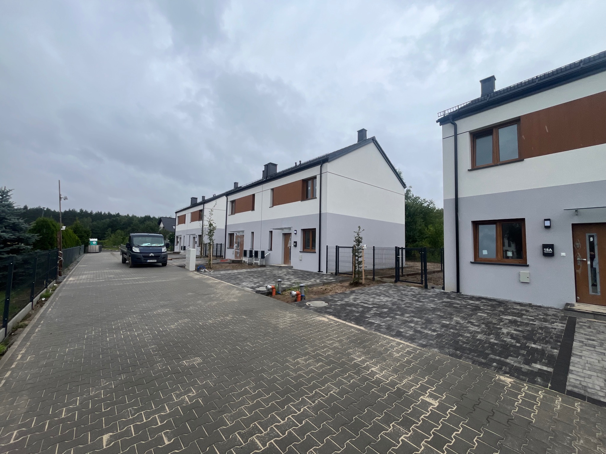 Szeregówka domów jednorodzinnych z elewacją w odcieniach bieli i szarości, z brązowymi akcentami, widok z ulicy wyłożonej kostką, pochmurne niebo.