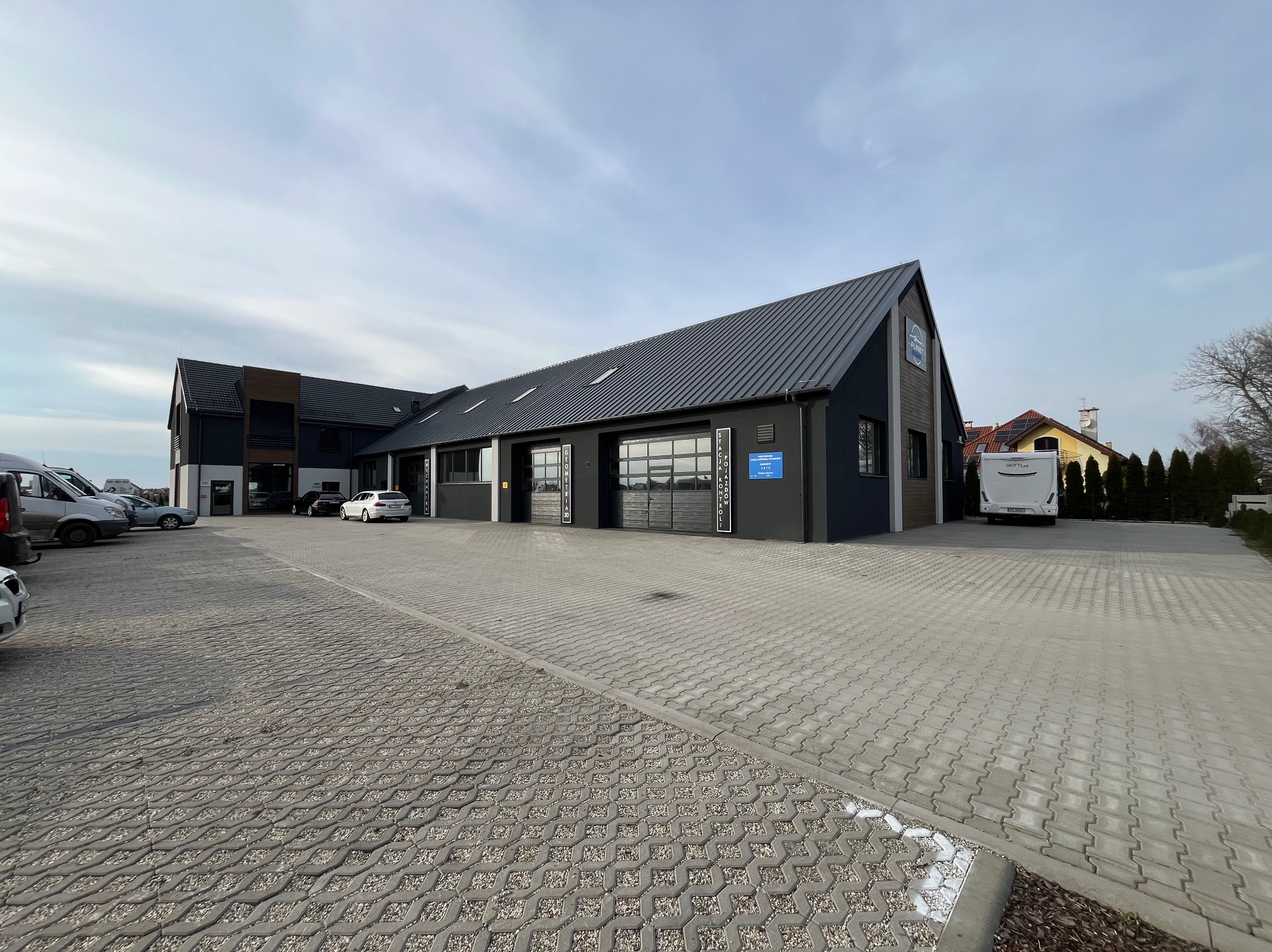 Nowoczesny budynek usługowy z ciemną elewacją i szarą kostką brukową na parkingu, widok z zewnątrz w pochmurny dzień.