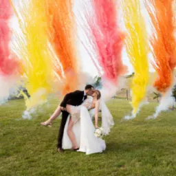 Para młoda w plenerze, mężczyzna trzyma kobietę w ramionach, w tle kolorowe pirotechniczne efekty specjalne w odcieniach żółci, pomarańczy i różu.