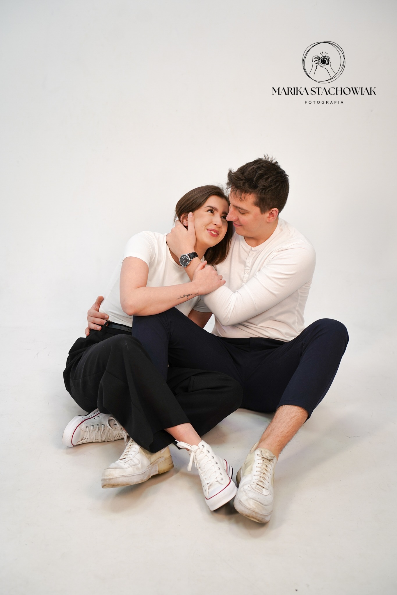 Zakochana para siedzi na białym tle, ona w białym t-shircie i czarnych spodniach, on w białej koszuli z długim rękawem, obejmują się i patrzą na siebie, widoczny zegarek na ręce kobiety i logo...