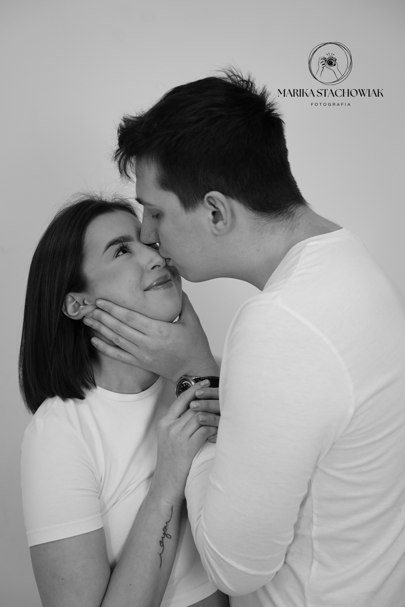 Czarno-białe zdjęcie narzeczonej z tatuażem na przedramieniu, delikatnie uśmiechniętej, gdy narzeczony całuje ją w czoło, dłońmi obejmuje jej twarz, na nadgarstku widoczny zegarek.