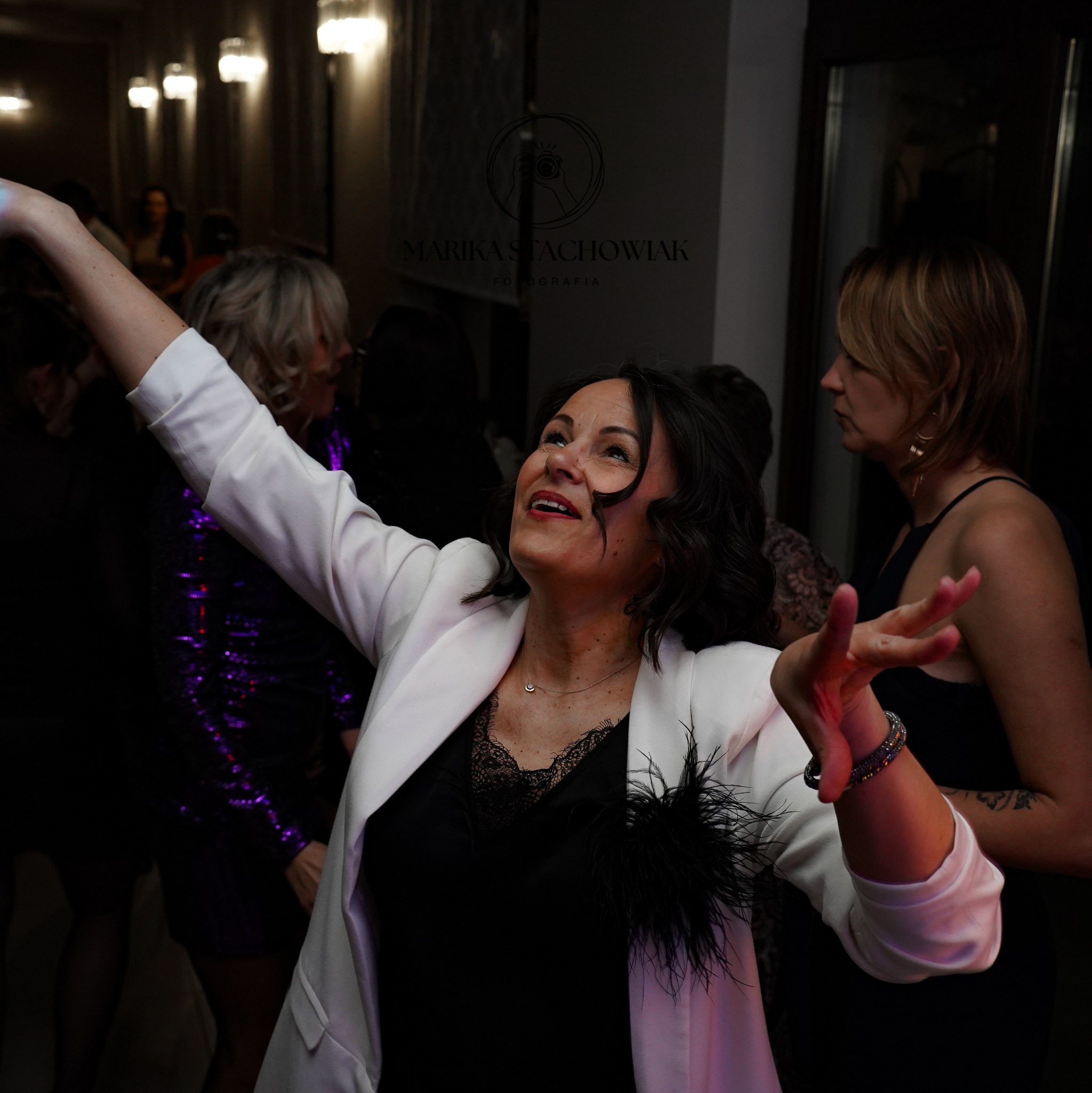 Uśmiechnięta kobieta w białej marynarce i czarnej bluzce z koronką, wznosi ręce do góry podczas eventu; w tle widoczni inni uczestnicy imprezy i dekoracyjne oświetlenie.