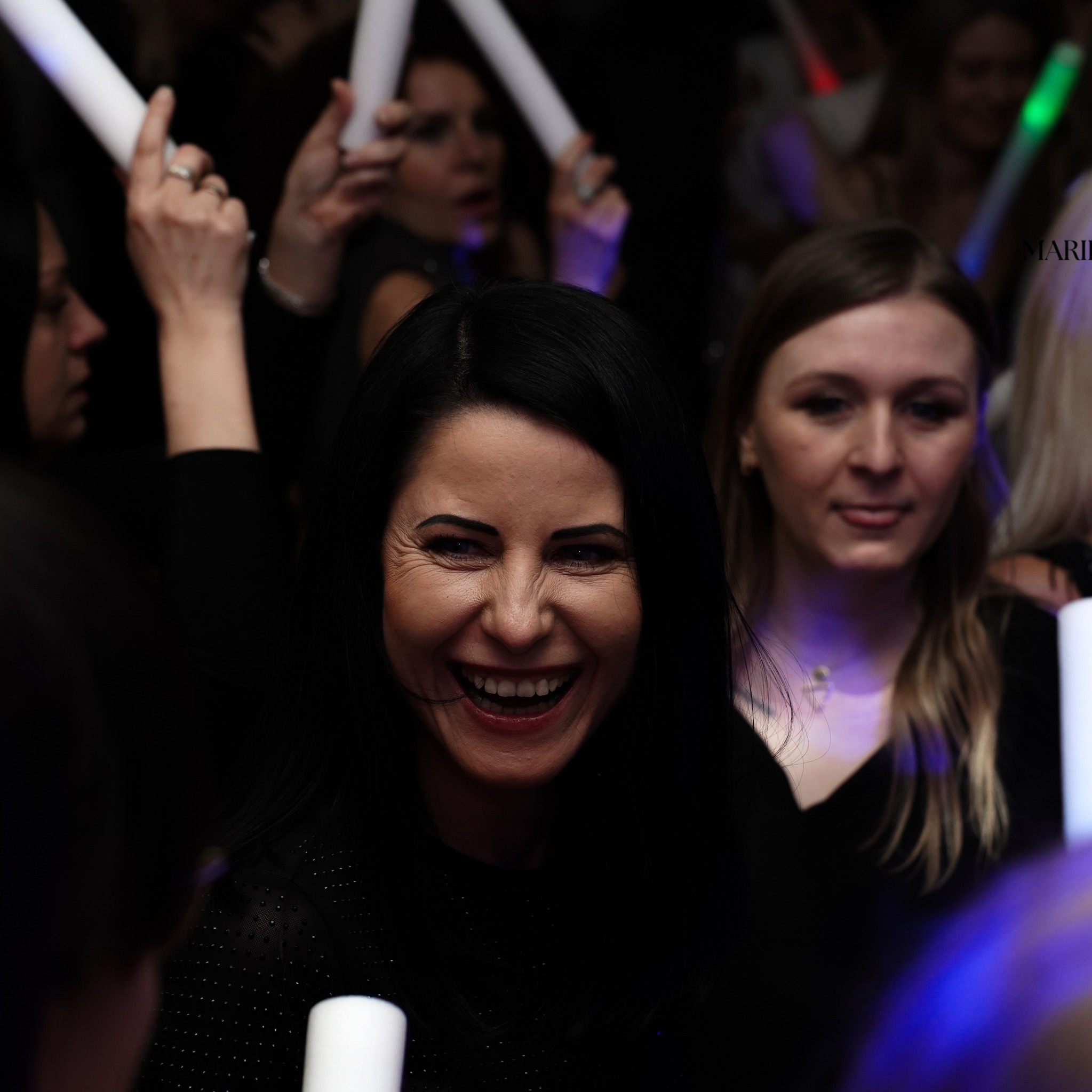 Rozbawiona kobieta z czarnymi włosami śmieje się na imprezie, w tle inne osoby trzymają świetlne pałeczki.