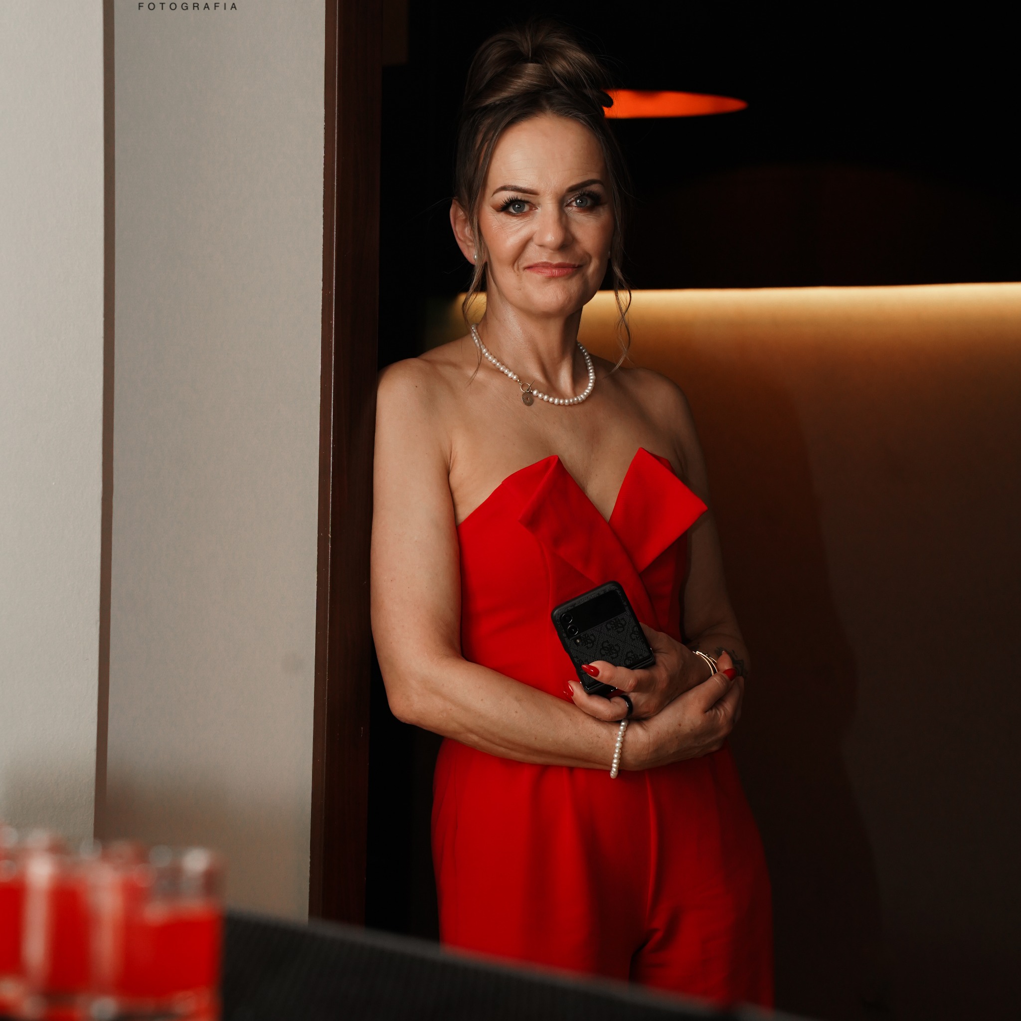 Elegancka kobieta w czerwonym kombinezonie bez ramiączek, z perłowym naszyjnikiem i bransoletką, trzyma czarny telefon z wzorem, stojąc przy ścianie z ciemnym tłem i rozmytymi szklankami...