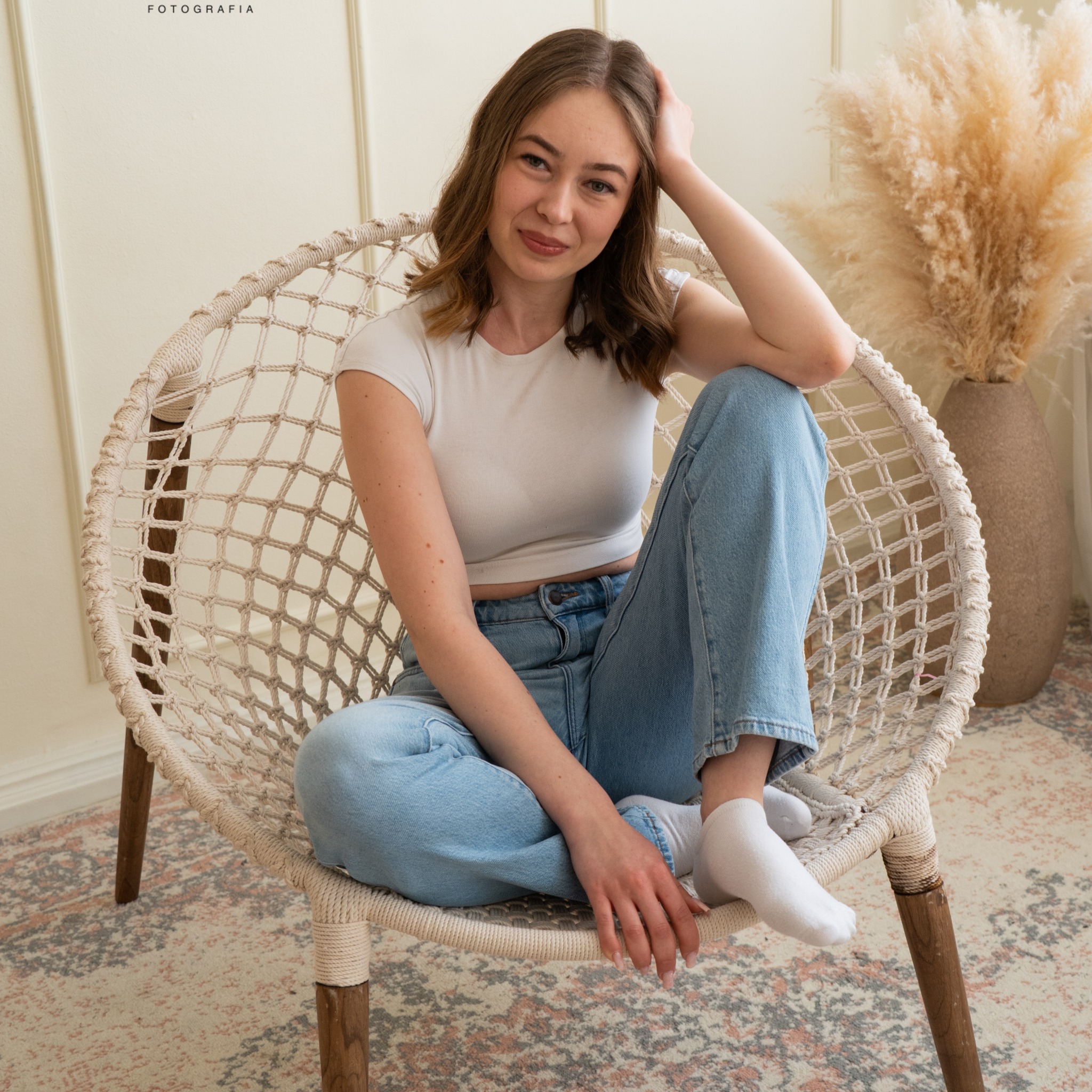 Portret młodej kobiety siedzącej w plecionym fotelu w jasnym wnętrzu z dekoracyjną trawą pampasową w tle.