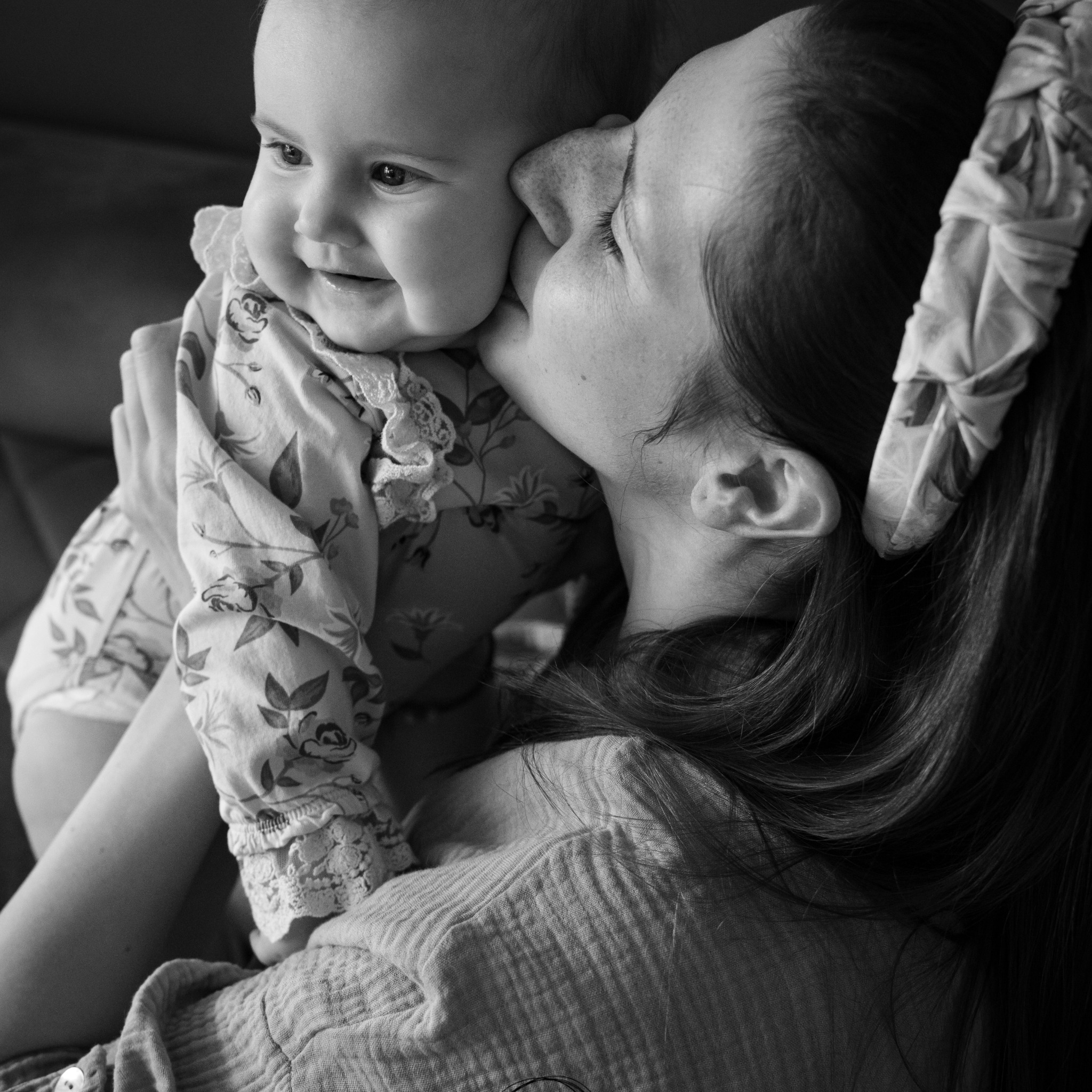 Czarno-białe zdjęcie matki całującej w policzek uśmiechnięte niemowlę ubrane w kwieciste body z koronkowym wykończeniem; kobieta ma na włosach wzorzystą opaskę.