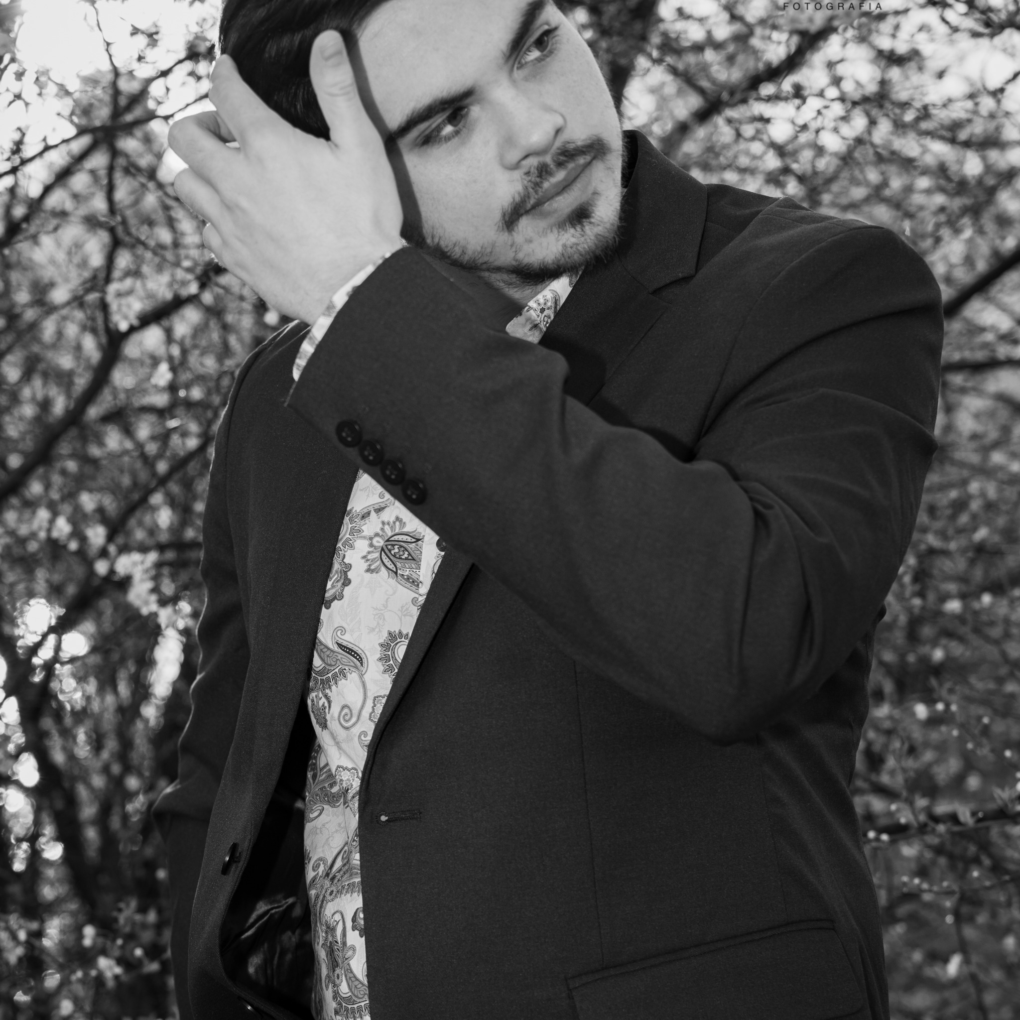 Czarno-białe portretowe ujęcie mężczyzny w garniturze i wzorzystej koszuli na tle drzew, z ręką uniesioną do włosów.