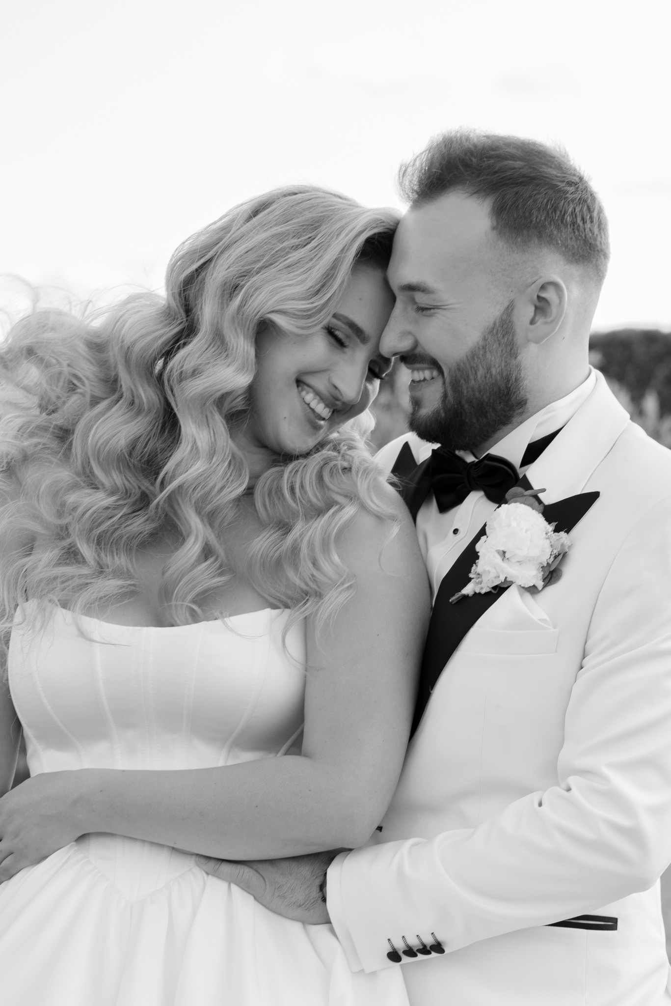 Czarno-białe, bliskie ujęcie uśmiechniętej pary młodej w objęciach. Ona w sukni z gorsetem, on w białym smokingu z muchą. Romantyczny nastrój.