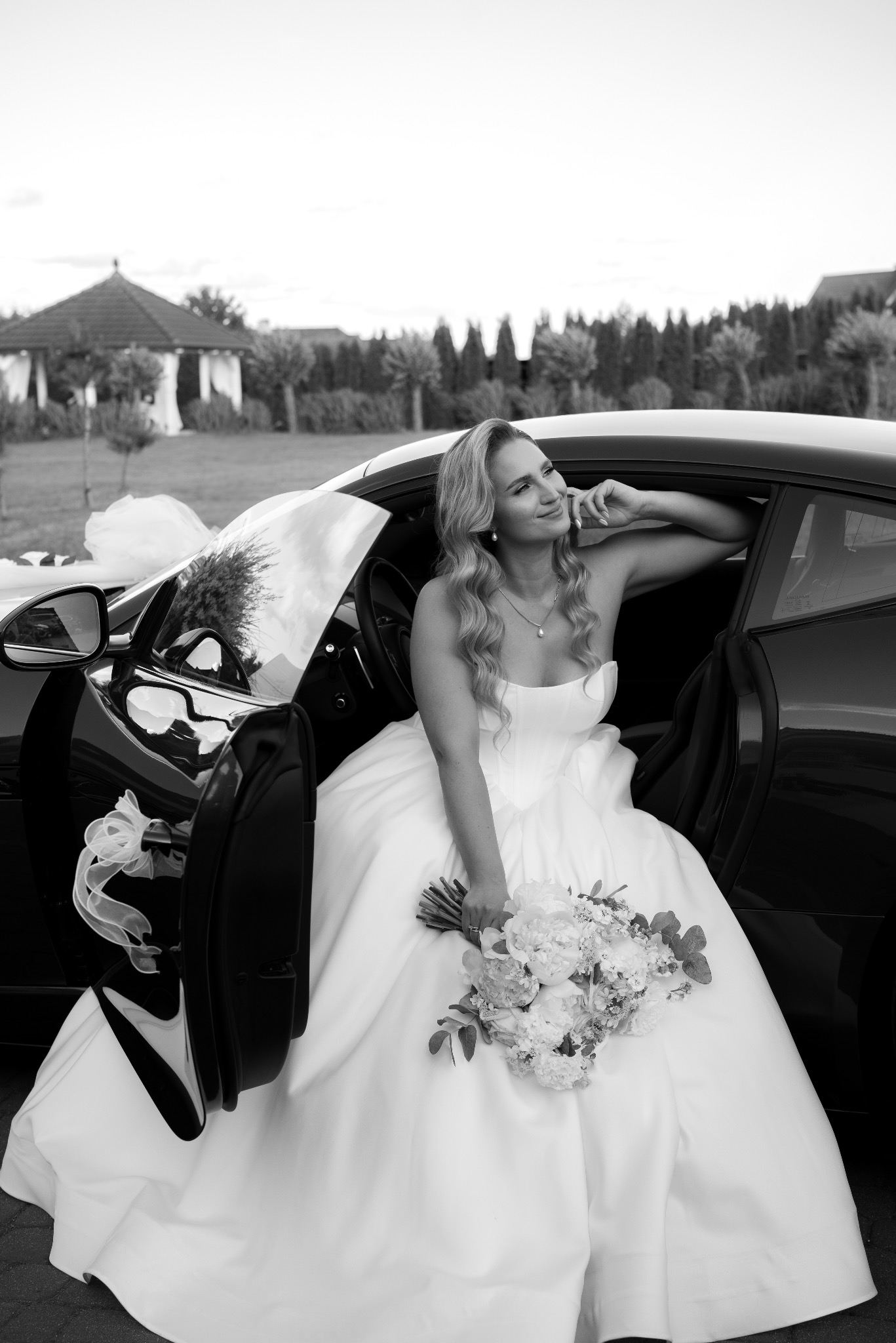 Elegancka panna młoda w sukni ślubnej, siedząca w czarnym samochodzie sportowym z bukietem, w tle altana i zieleń. Ujęcie w czerni i bieli.