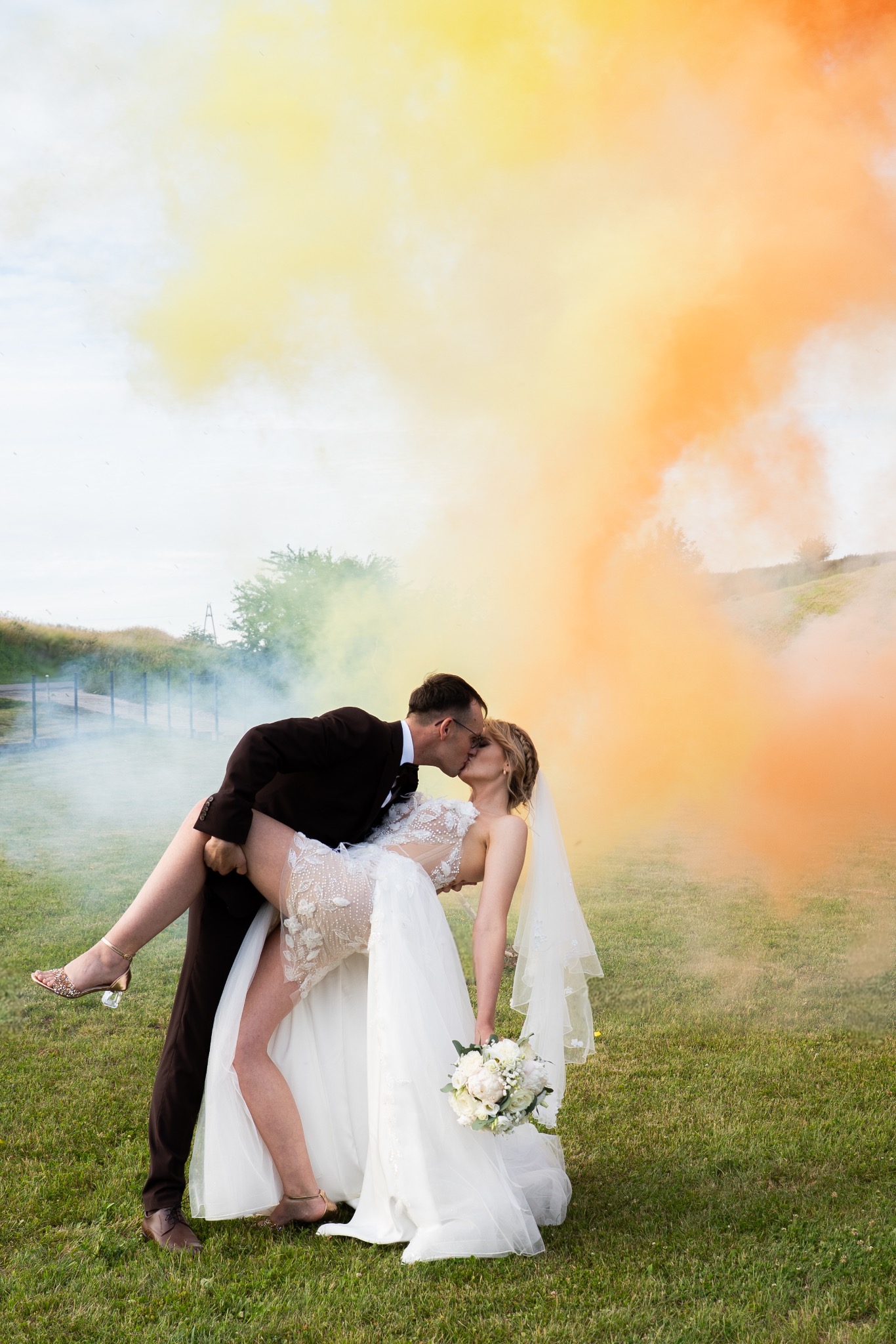 Para młoda w tańcu, otoczona kolorowym dymem. Panna młoda w koronkowej sukni i welonie, trzyma bukiet. Pan młody w brązowym garniturze, unosi ją w ramionach. Romantyczna sceneria.